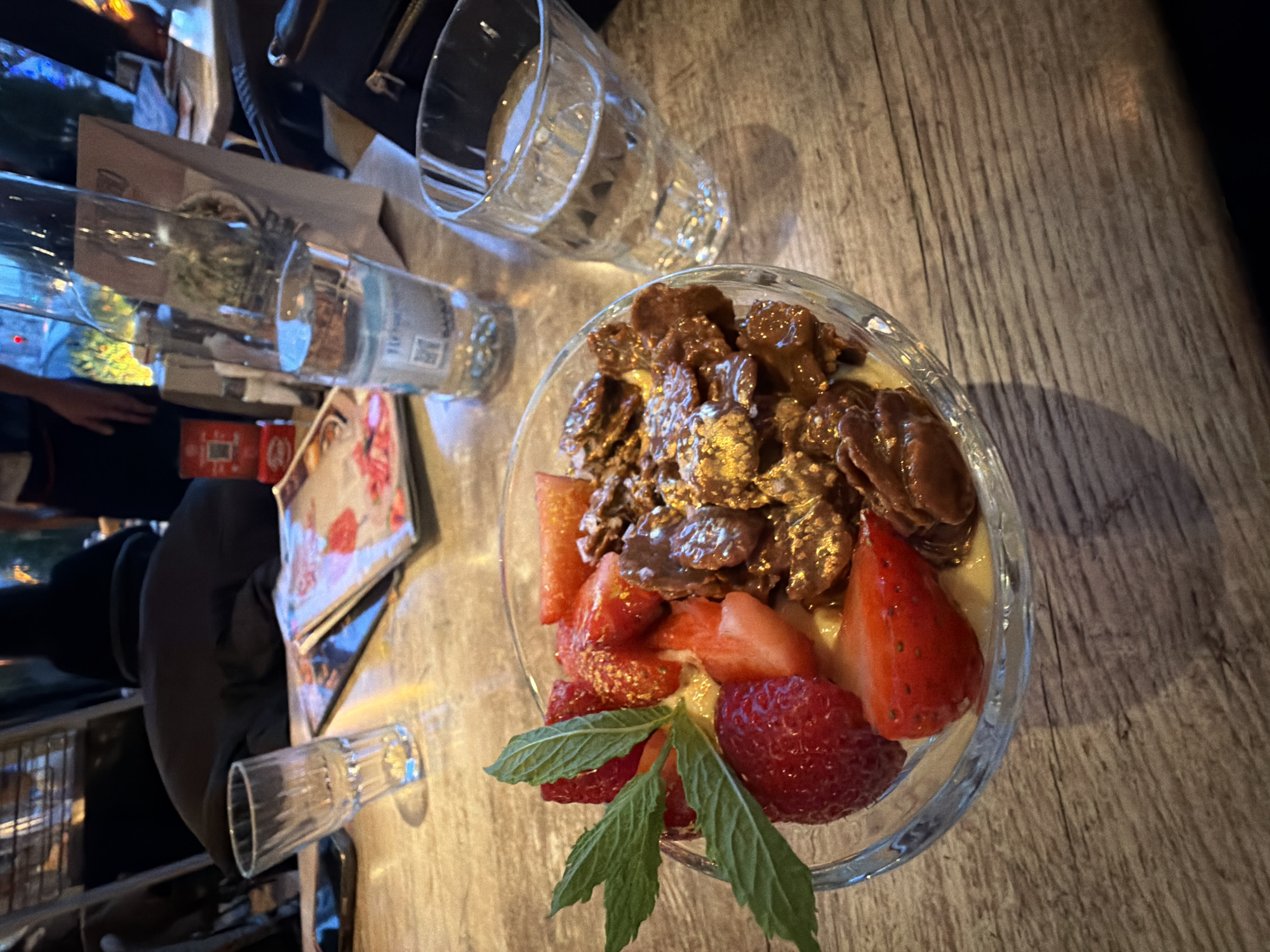 An overhead shot captures a glass bowl filled with dessert, prominently displayed on a wooden table. The bowl contains a light-colored base, likely yogurt or pudding, topped with chocolate-covered flakes or cereal pieces. Fresh strawberries, some halved and some quartered, are arranged around the edge of the bowl, with two green mint leaves serving as a garnish.
On the table, several other items are visible: two clear drinking glasses (one empty and faceted, another plain glass containing liquid, possibly water), a small empty shot glass, and a clear water bottle. The label on the water bottle partially shows "330 ML". Menus or paper items, along with a red box and a black bag, are seen in the blurred background.
The setting appears to be an indoor cafe or restaurant in Sofia, Bulgaria, indicated by the table, glassware, and prepared food. The lighting is subdued, suggesting evening or artificial illumination, with shadows cast by the objects. A person's hand is partially visible on the left side of the frame, and a dark-clad figure is vaguely discernible in the background, implying a dining or social activity. The image has a warm, inviting tone.