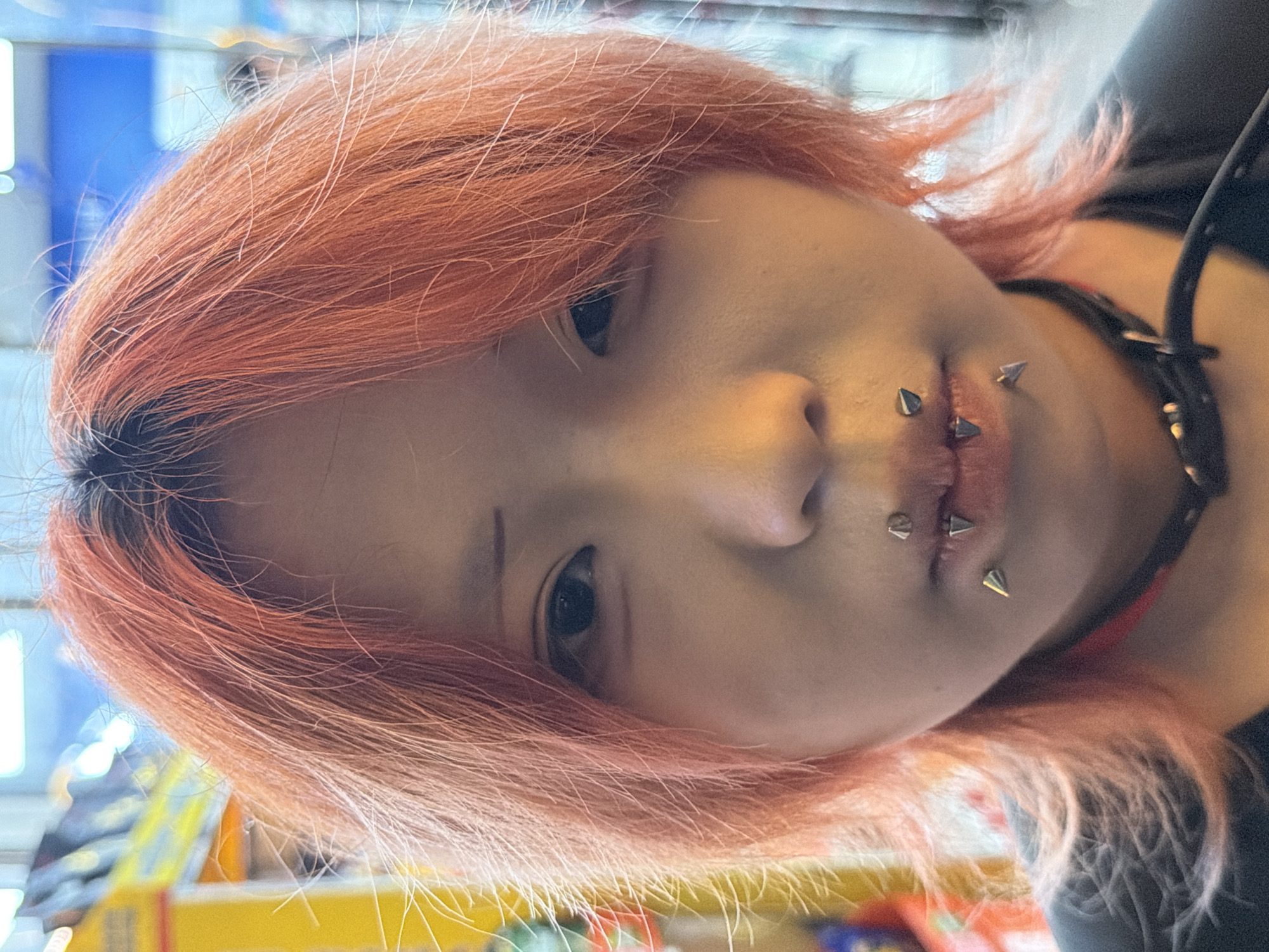 A headshot captures a young individual with short, choppy, reddish-orange hair featuring visible dark roots. The person has a pale complexion, dark eyes, and a neutral expression while looking directly at the viewer. Multiple metallic spike-shaped piercings are visible: two below the lower lip, one on the right side of the lower lip (viewer's left), and a smaller stud or spike above the upper lip. A black choker adorned with small metallic studs is worn around the neck. The background is out of focus, showing blurred areas of yellow, blue, and white, indicative of an indoor setting with artificial lighting, possibly a commercial establishment. The scene is located in Jiangbeicheng Subdistrict, China.