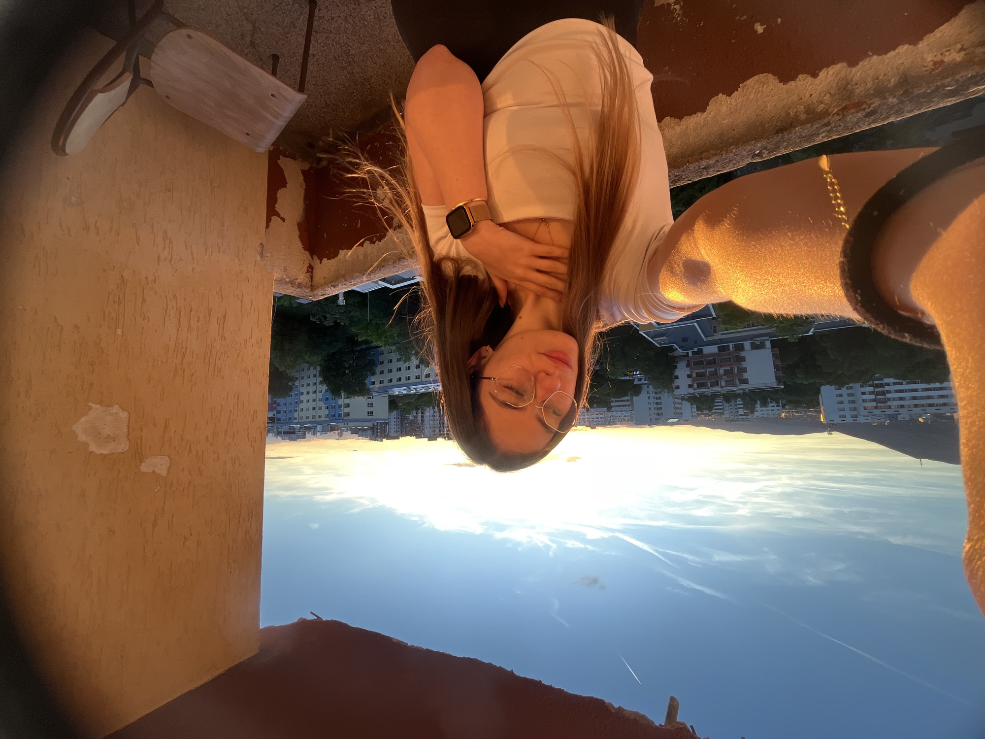 A young woman with long brown hair and glasses takes a selfie on a balcony overlooking a city at sunset. She is wearing a white t-shirt and a black skirt. The woman has her right hand on her chest and her left arm is extended, holding the camera. The sun is setting behind a cityscape, casting a warm golden light on the scene. The balcony railing is made of concrete and appears weathered. A single chair with a wooden seat and metal legs is visible on the balcony. The sky is a clear blue with some wispy clouds.