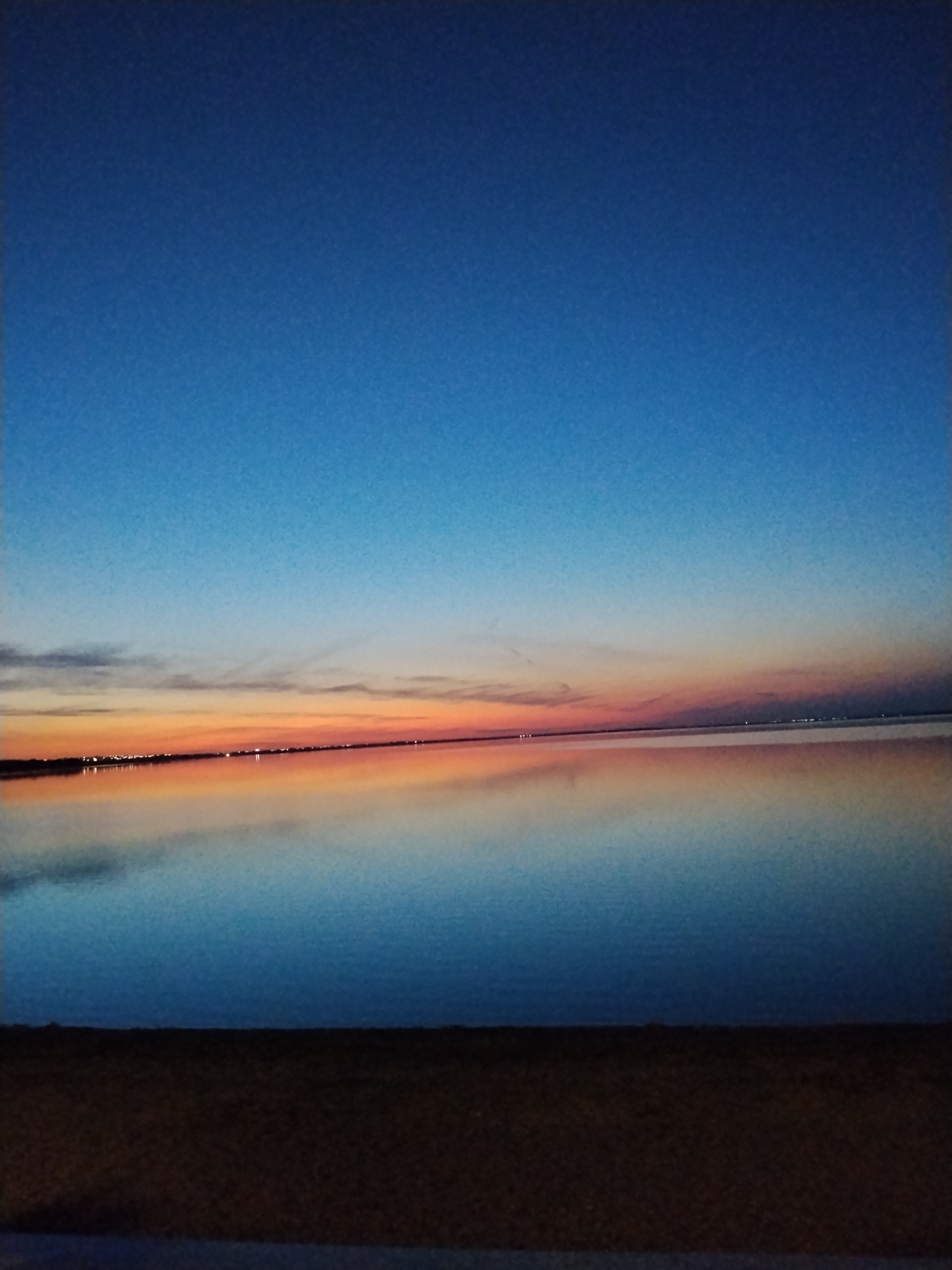 The image depicts a serene sunset over a body of water, likely a lake or sea, with a silhouetted shoreline and a strip of dark ground in the foreground. The sky transitions from a deep blue at the top to lighter shades of blue, pink, and orange near the horizon, reflecting the vibrant colors of the sunset onto the calm water. Small, distant lights are visible along the shoreline, suggesting a town or city. The overall scene evokes a sense of tranquility and the natural beauty of the twilight hour. The location is identified as Kokshetau, Kazakhstan, suggesting the image was taken near the city.