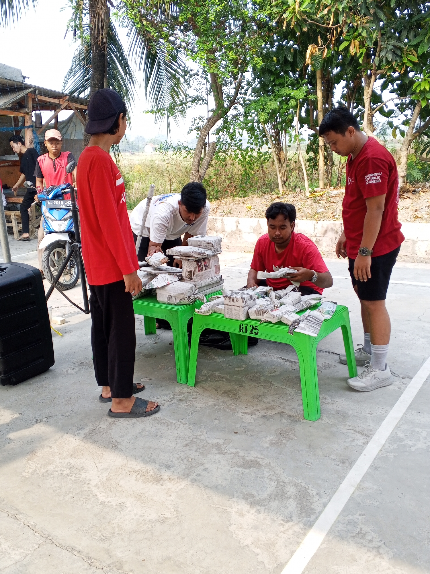 A group of five young men are gathered around a bright green plastic table outdoors. The scene appears to be in a semi-rural area in Ciruas, Indonesia, with trees and vegetation in the background, and a paved ground surface marked with white lines, suggesting it might be a sports court.
Three of the men are wearing red shirts, and two are wearing darker colored clothing. One man in a red shirt, seated, is sorting through packages wrapped in newspaper. He is wearing a watch. Another man in a red shirt, standing behind the table, is wearing black shorts and grey socks and sneakers. The third man in a red shirt, to the left, is wearing black pants and dark sandals, and is looking away from the table. Two other men are partially visible in the background. One is wearing a black shirt and dark pants, and the other is wearing a red shirt and a light-colored cap.
The table is covered with numerous packages, all wrapped in newspaper. They are arranged in stacks and rows, some appearing to be of similar size and shape. A blue and white motorcycle is parked to the left of the table. A large black speaker is also positioned to the left of the table. The lighting suggests it is daytime, with sunlight casting shadows. The overall impression is of a casual outdoor gathering, possibly related to a sale or distribution of items wrapped in newspapers.