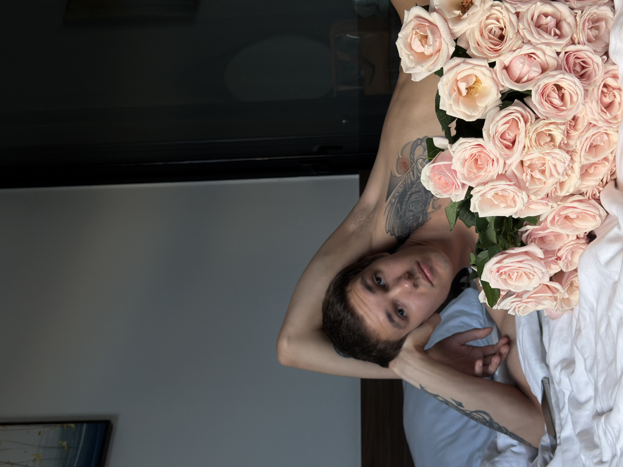 A young man with a tattoo on his chest and arm lies on a bed covered with white sheets. He is propped up on his left arm, with his head resting on his hand. In front of him is a large bouquet of pale pink roses. The room appears to be a bedroom, with a headboard visible behind him and a doorway or window with dark glass to the right. There is also a framed picture on the wall in the upper left corner of the image. The lighting suggests it is daytime, possibly with natural light filtering in. The overall scene conveys a relaxed and intimate atmosphere. The provided context places this image in Đà Nẵng, Vietnam.