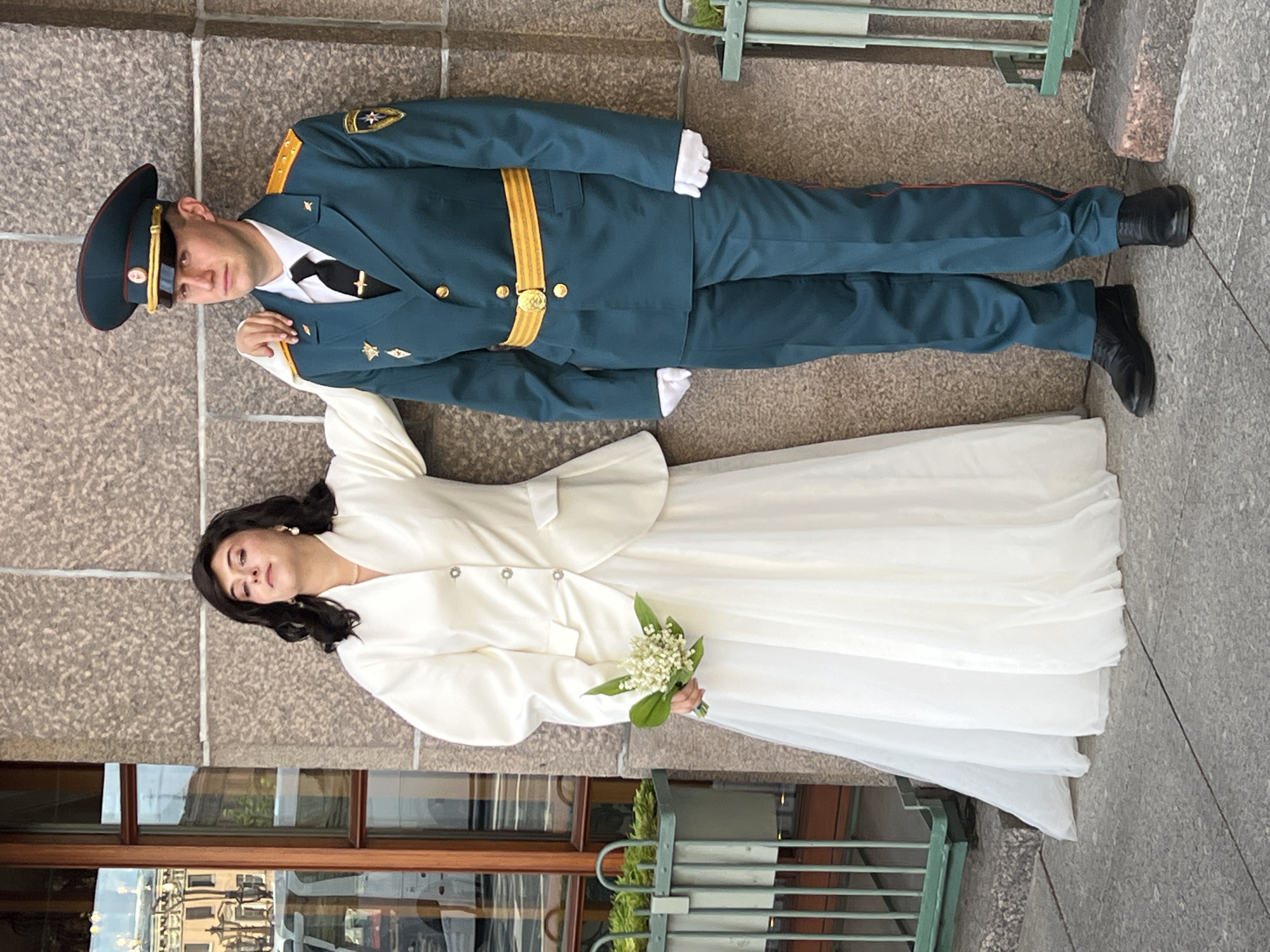 A man in a military uniform and a woman in a wedding dress pose for a photograph near a building, which appears to be located in Saint Petersburg, Russia based on its architectural style. The man is wearing a dark green military uniform with gold detailing, a black cap, and black shoes. The woman is wearing a white wedding dress and holding a bouquet of lilies of the valley. They are standing on a stone surface, next to the building's facade, which has a window reflecting a building across the street and a decorative railing.