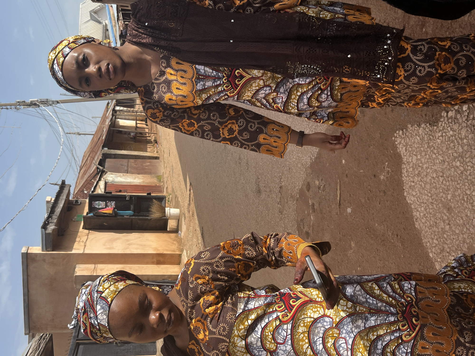 Two young women are standing in a dusty street lined with shops in Jos, Nigeria. Both are wearing vibrantly patterned traditional clothing, with the woman on the left in a brown and gold outfit with a colorful headscarf, and the woman on the right in a similar outfit with a dark brown lace shawl. The woman on the left is looking down at her phone, which she is holding in her right hand. The woman on the right is looking directly at the camera. The buildings in the background appear to be simple shops with corrugated metal roofs, typical of the area. The ground is a mix of packed dirt and small stones. The sky is clear and blue, indicating a sunny day. The scene conveys a sense of everyday life in an urban Nigerian setting.