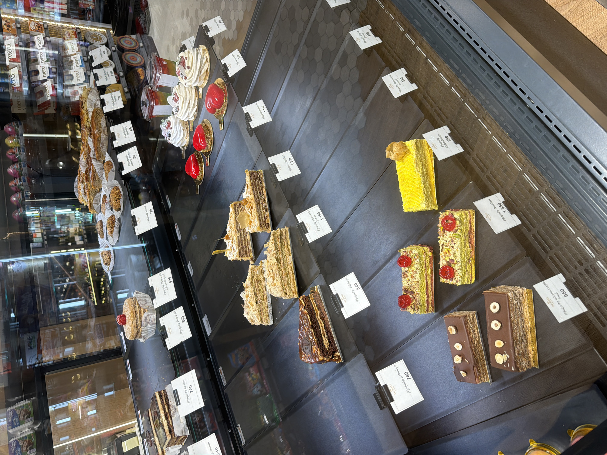 The image shows a vibrant, well-lit display case filled with an assortment of pastries and cakes, likely in a bakery or supermarket setting in Yerevan, Armenia. The display is arranged on multiple tiered shelves within a refrigerated unit, presenting a wide selection of confectionery.
Various types of pastries are visible. On the upper shelves, there are individual round desserts topped with whipped cream and berries, alongside glossy red, heart-shaped cakes. Further down, there are numerous sliced cakes with multiple layers, featuring creamy fillings, nuts, and different toppings. These include cakes with visible layers of sponge and cream, some coated in chocolate and adorned with hazelnuts, others with a light, crumbly texture. There's a notable bright yellow cake, and green, pistachio-like cakes topped with red raspberries.
Each pastry has a small white price tag next to it, displaying prices in Armenian Dram (AMD) and names of the pastries in Armenian. "SAS SWEET" is printed on many tags. Armenian text visible includes "Թխվածք" (Tkhvatsk - cake/pastry) followed by descriptions such as "մեղրային" (honey), "անտառային" (forest), "Կամիլա", "սնիկերս" (Snickers), "տղամարդու իդեալ" (man's ideal), and "Մանն մանգո". Prices range from 380 AMD to 1350 AMD.
In the background, beyond the main pastry display, shelves are stocked with packaged goods, most notably multiple boxes of "Merci" brand chocolates. Further back, reflections in the glass hint at another display unit containing bottles, possibly wine or spirits. The visible floor has a grey hexagonal tile pattern. The scene captures a moment of a retail environment, showcasing an appealing array of sweet treats.