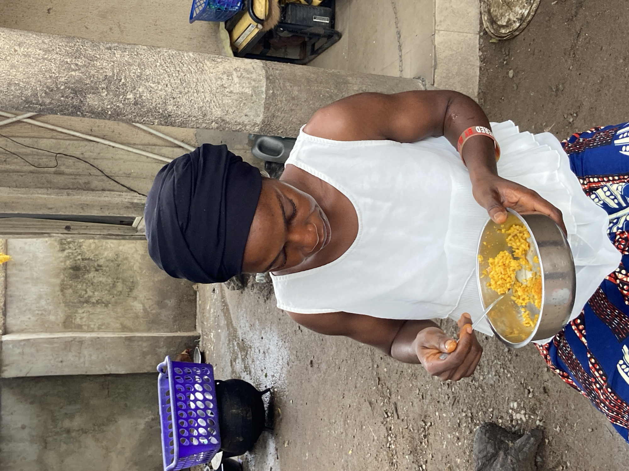 In an outdoor setting in Mararaba, Nigeria, a woman wearing a dark head wrap and a white top over a blue patterned skirt is holding a metal bowl of cooked yellow rice and a spoon. She appears to be preparing or eating the meal. The ground around her is concrete. Behind her, there is a concrete structure and a blue plastic crate. A black cooking pot is also visible near the crate.