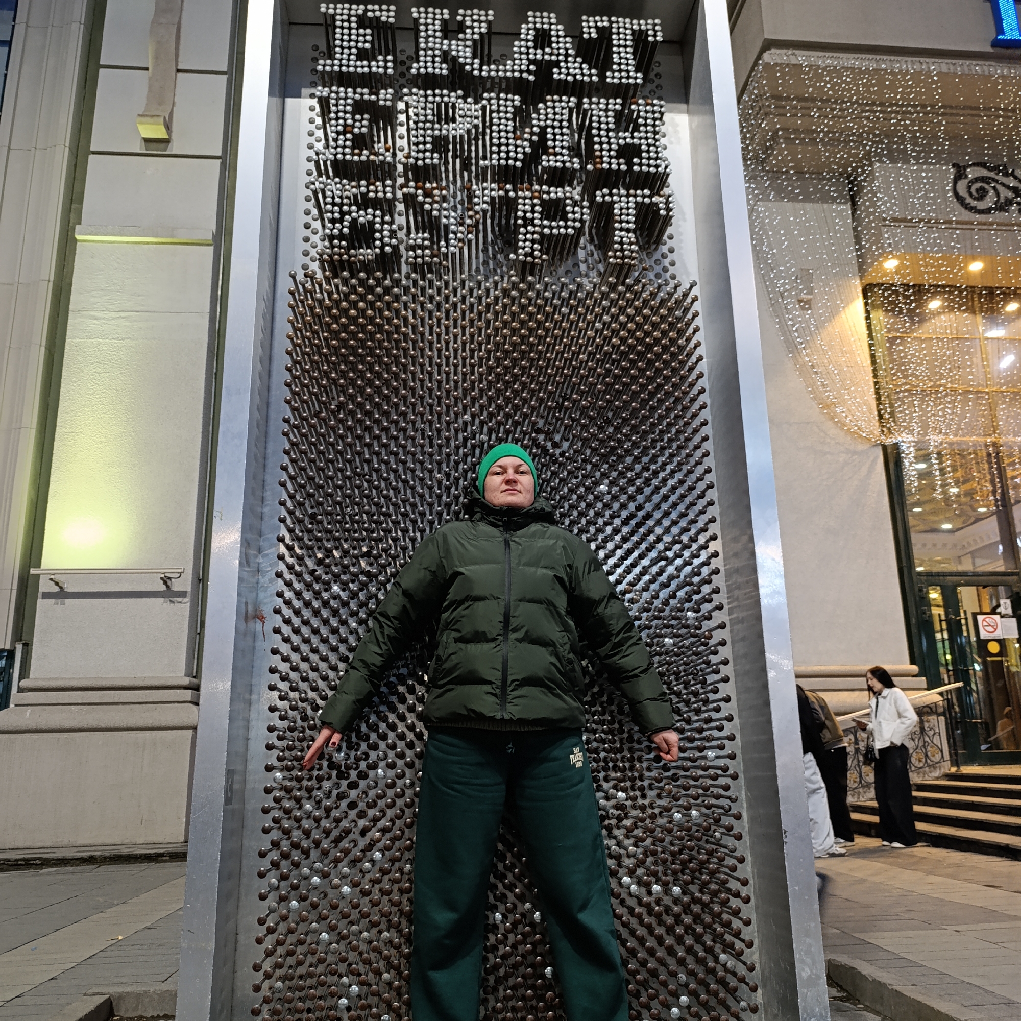 The image depicts a person interacting with a large, vertical pin art display in an urban environment at night or dusk, indicated by the artificial lighting. The main subject, wearing a dark green puffer jacket, matching dark green pants with "SAN FRANCISC 1900" printed on the left thigh, and a green beanie, stands pressed against the metallic pins of the display. Their arms are slightly outstretched, and their hands, with visible red nail polish, are also pressed into the pins. They have a neutral expression.
The prominent pin art installation is made of numerous metallic pins with rounded heads, a mix of silver and bronze tones. The upper section of the display clearly forms the word "EKATERINBURG" in block letters using the protruding silver pins. Below this text, the pins are pushed inward by the subject's body.
In the background, to the right of the pin art display, a building facade is brightly lit and adorned with numerous small, sparkling white lights, suggesting festive or commercial lighting. Another person in a white jacket and dark pants is visible further back, walking up a set of outdoor stairs. To the left, another building with light-colored, textured walls and architectural features is partially visible, illuminated by a warm, yellow upward-shining light. The ground is paved, suggesting a sidewalk or public area in the city of Yekaterinburg, Russia.
