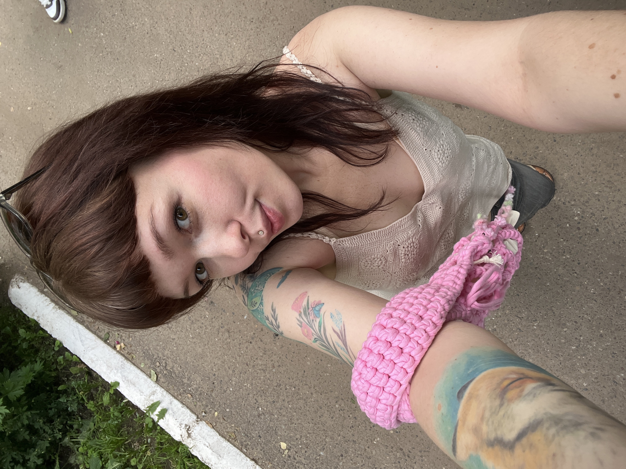 In Chaikovsky, Russia, a fair-skinned woman with shoulder-length brown hair, bangs, and light green eyes is captured in a selfie. She has a lip piercing. Her left arm displays colorful tattoos, including floral designs and a landscape. She is wearing a light-colored, crocheted tank top and dark jeans. A bright pink crocheted accessory is wrapped around her left forearm. She is standing on a paved surface with a white concrete curb and green foliage to her left. In the background, a shoe can be seen on the pavement.