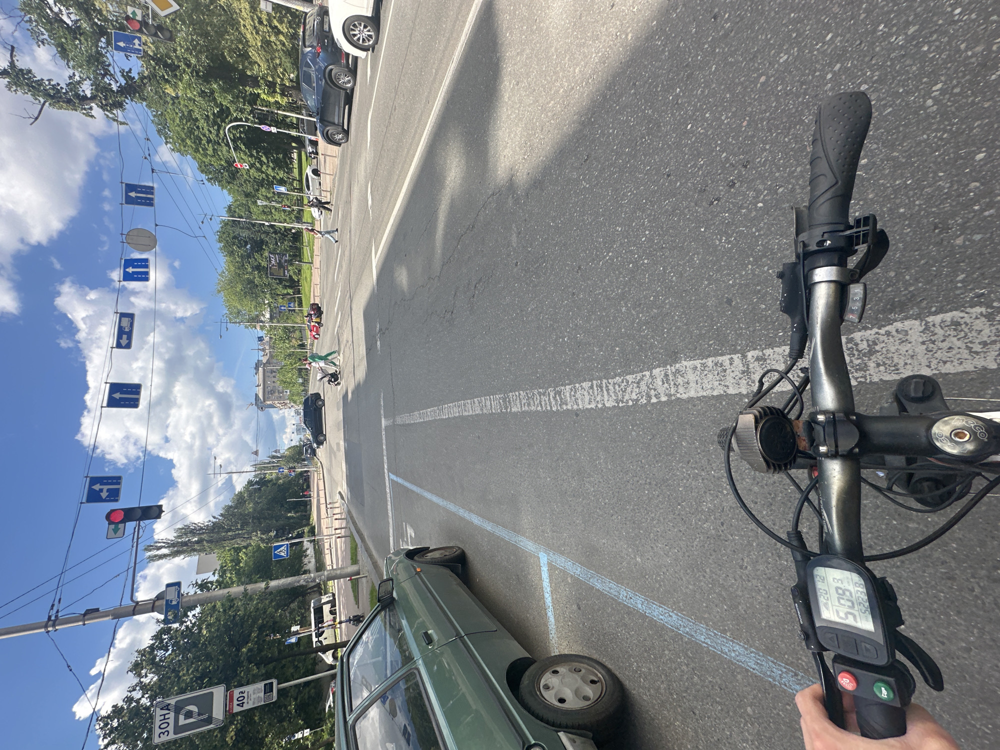 This media file is a first-person perspective video shot from a bicycle, likely in Kyiv, Ukraine. The scene is an urban street on a sunny day, with blue skies and fluffy white clouds visible. The camera is oriented vertically, giving a dynamic view of the surroundings.
The main subject is the bicycle's handlebars and front wheel, with a digital display showing speed, battery level, and other cycling data. The display shows "20 12" and "50.9" and "5223.8". There are also two buttons on the handlebars, one red and one green.
The setting is a road with marked lanes and a sidewalk. Several cars are parked or driving on the road, including a light green car in the foreground and a dark car further down the street. Pedestrians are visible on the sidewalk, some crossing the street. Traffic lights and various road signs are present, including directional signs and speed limit signs. One sign shows "40" with a circle around it, indicating a speed limit of 40. Another sign shows "30HA" and a parking symbol.
The activity taking place is cycling through the city. The sunlight suggests it is daytime, likely mid-morning or afternoon. The overall mood appears to be ordinary urban travel.
