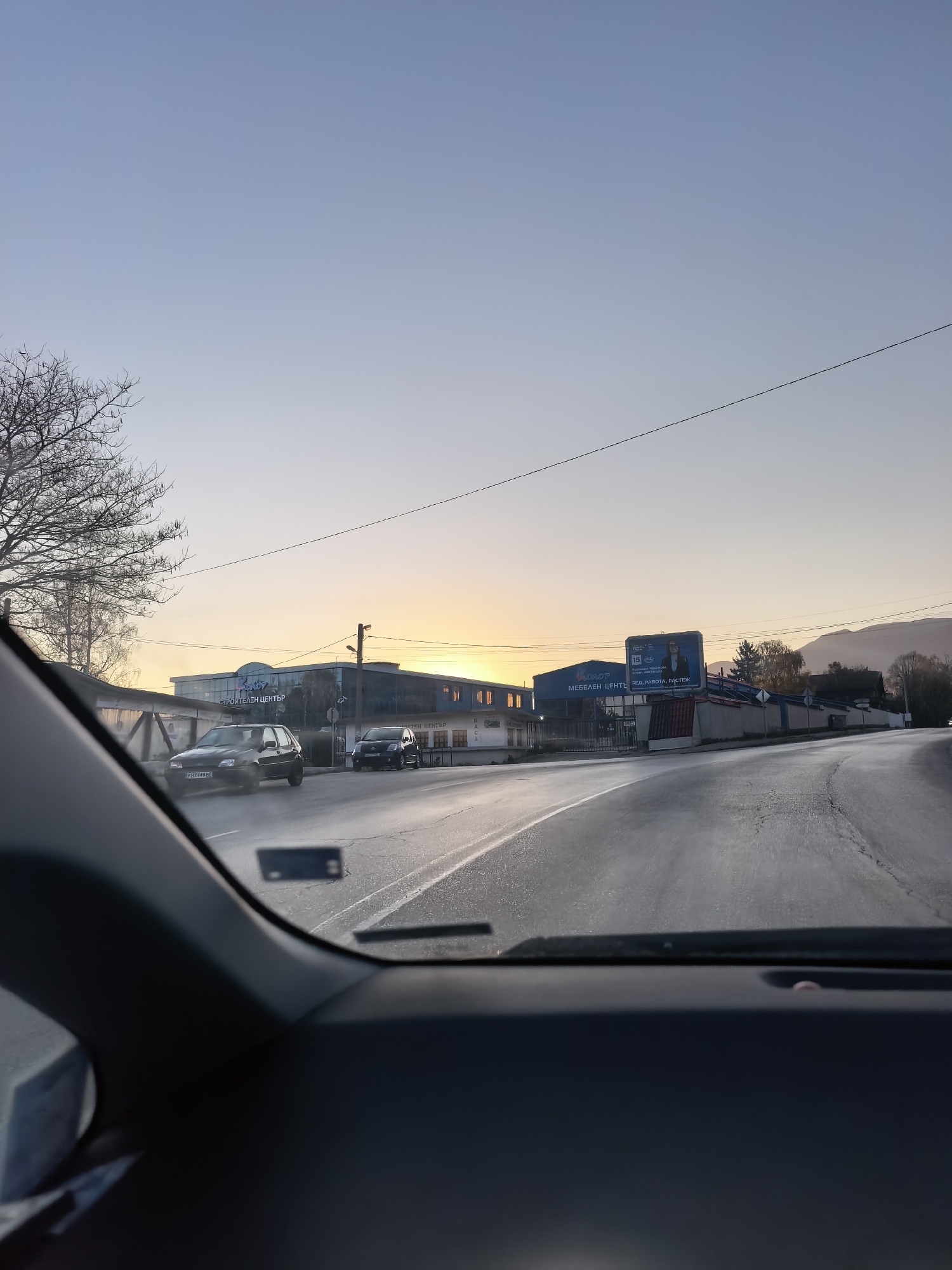 The image depicts a view from inside a car driving on a wet road in what appears to be a town or city. The sky is a pale blue, indicating either early morning or late afternoon. In the mid-ground, there are several low-rise buildings. One building features the words "Строителен Център" and another has the word "КОЛОR" along with Bulgarian text. A billboard promotes furniture and states "МЕБЕЛЕН ЦЕНТР РЕД, РАБОТА, РАСТЕЖ". Cars are visible on the road, and some trees can be seen on the left side of the frame. The environment suggests a location in the city of Unknown, Unknown.