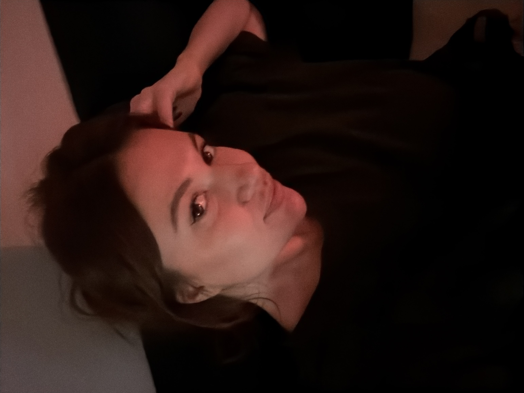 A medium shot shows a woman reclining with her head resting on a surface with a dark background. The light is dim and red, casting a warm hue on her face. She is wearing a dark-colored garment. Her left hand is near her head. Due to the low light and limited visual information, the location within Belgrade, Serbia, cannot be determined.