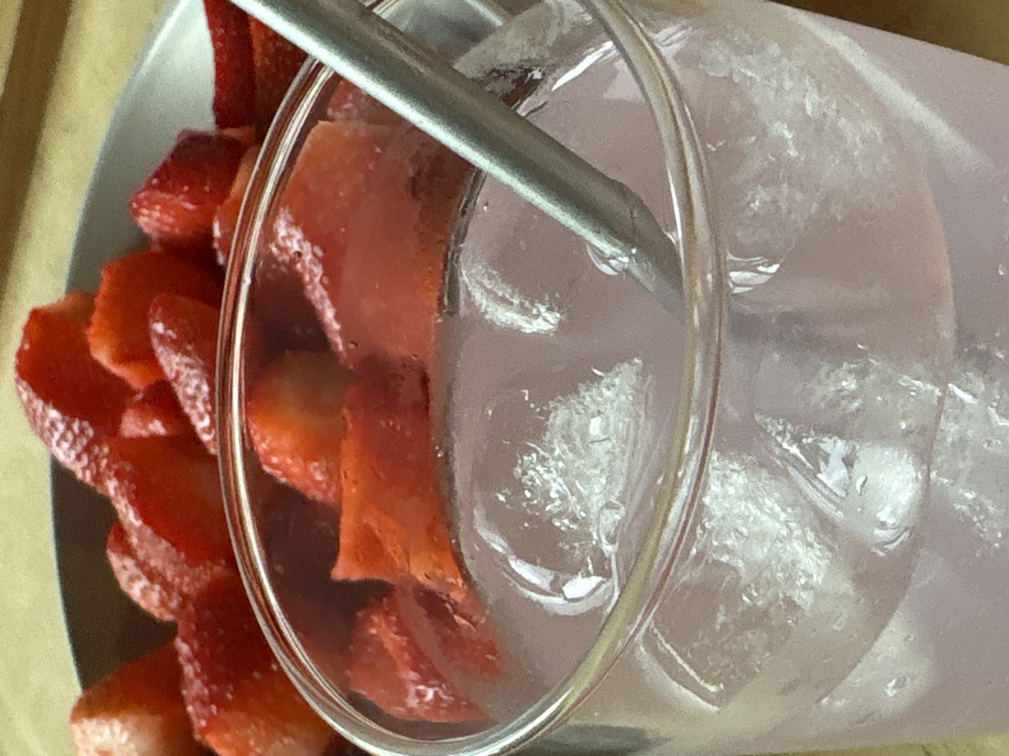 The media file captures a close-up, top-down view of a refreshing drink accompanied by fresh fruit. The central object is a clear glass, filled with a pale purple or pink liquid and numerous ice cubes. A slender, silver metallic straw extends into the drink from the top left. To the left of the glass, a white plate or shallow bowl holds a generous serving of bright red, sliced strawberries. The strawberries appear fresh, cut into various pieces, such as halves and quarters, with some juice visible on their surfaces. The liquid in the glass exhibits reflections and refractions from the ice and the glass's rim. The background is a soft, textured surface, likely wood, in shades of beige and light brown. No people are visible, nor is any text. The image suggests a moment of enjoyment or a prepared beverage with a healthy fruit side.