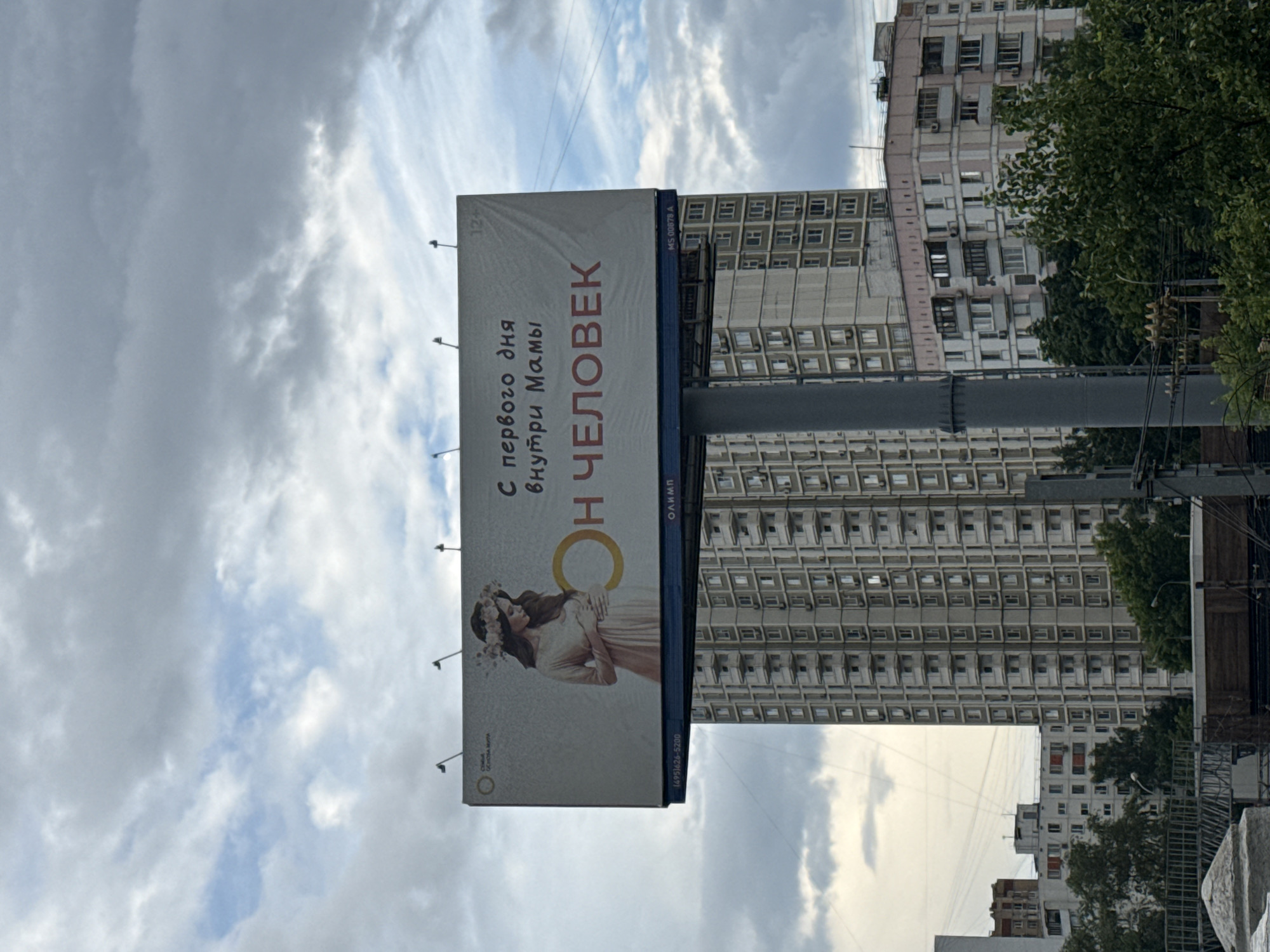This is a vertical, eye-level shot of a billboard in Moscow, Russia. The billboard features a pregnant woman with a flower crown, suggesting a theme of new life or motherhood. Text on the billboard is in Russian and translates to "From the first day inside Mom, HE IS HUMAN." A phone number "(495)626-5200" and "OLYMP" are also visible. The billboard is positioned in front of a dense urban background of tall, Soviet-era apartment buildings. The sky is overcast with dramatic clouds, indicating a potentially moody or transitional weather condition, possibly late afternoon or early evening. The overall scene is one of urban life juxtaposed with a message celebrating human development.