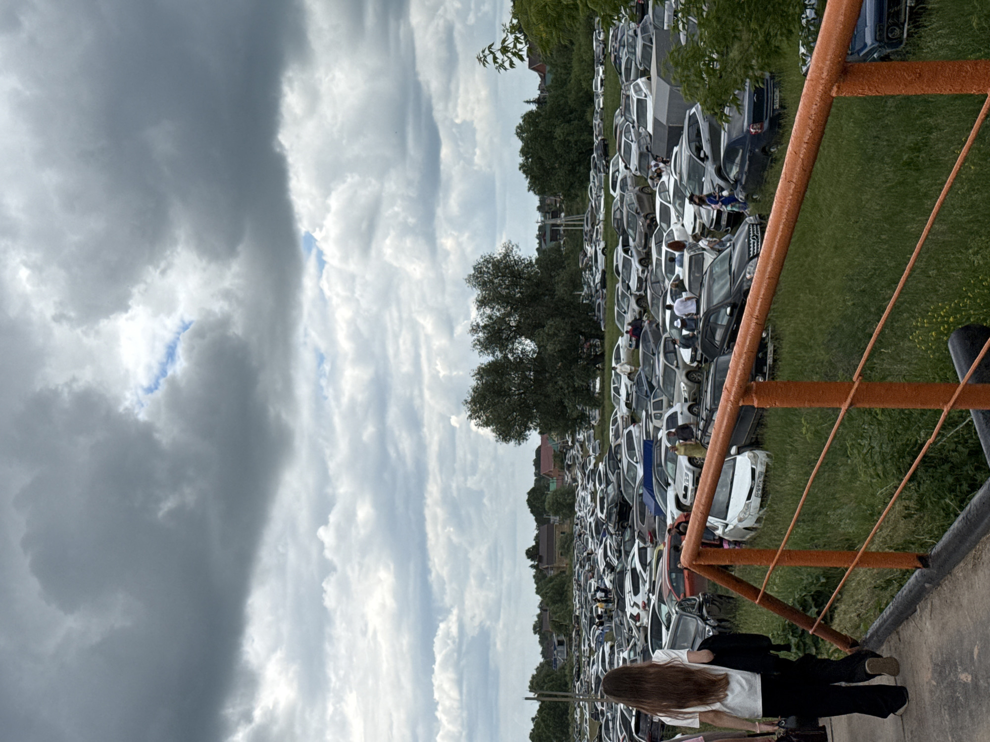 The image depicts a large, outdoor parking lot filled with numerous vehicles, predominantly cars. The parking lot appears to be situated in a semi-rural or suburban area, with trees and some buildings visible in the background, suggesting it might be near the city of Nurkeevo, Russia. The sky is overcast with heavy clouds. In the foreground, a person with long brown hair, wearing a white t-shirt and black pants, is walking away from the camera. The person is carrying a black bag. An orange railing runs across the lower portion of the image, separating the foreground from the parking lot. Several other individuals are scattered throughout the parking lot, some standing near vehicles, and others appearing to be walking. The ground in the parking lot is a mixture of grass and dirt, with some vehicles parked on grassy areas.