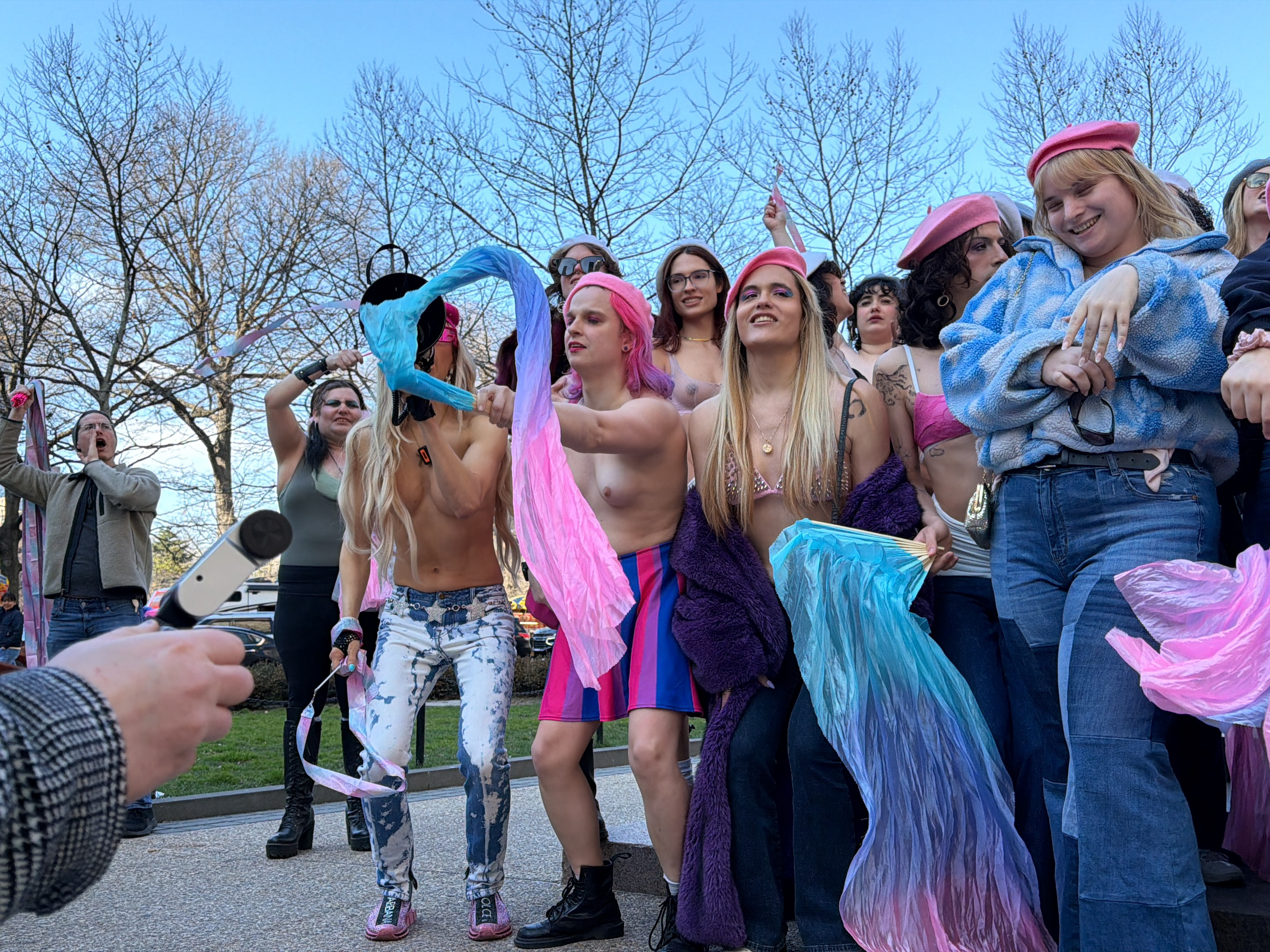 A group of people are gathered outside under a clear sky with bare trees in the background. Most of the individuals are young and fair-skinned. Some are topless. One person wears a blue-and-pink striped skirt, another wears acid-washed jeans, and several have pink berets. The group is holding and waving iridescent scarves. A person in the foreground is holding a device that may be a microphone or camera. The location is most likely not the city of New York, United States.