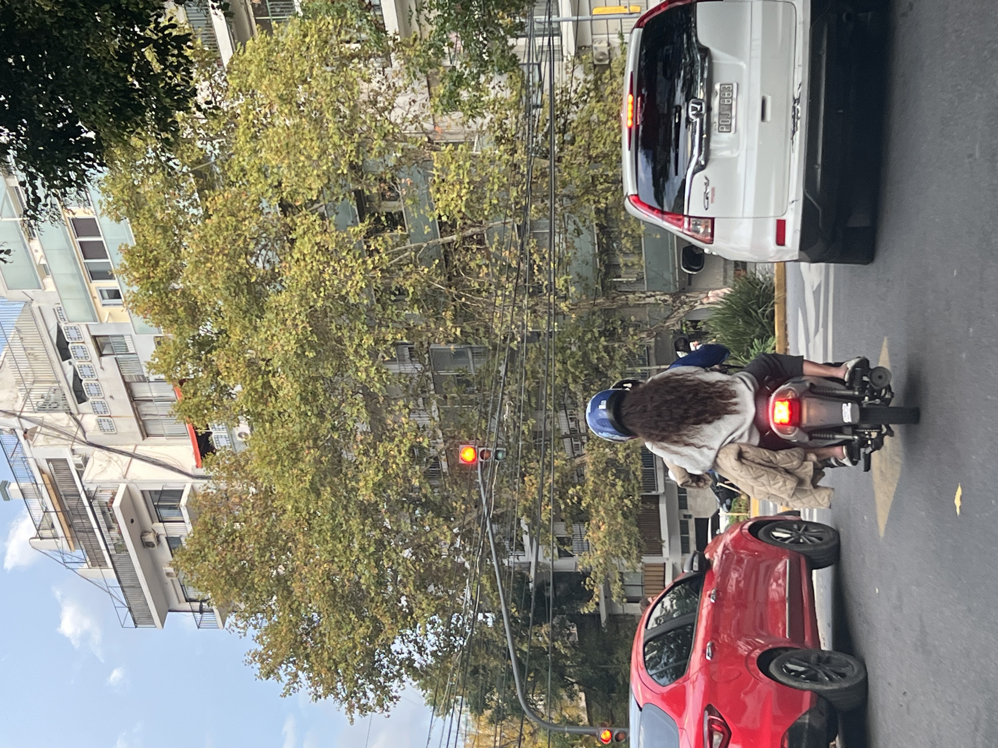 The image captures a street scene in Buenos Aires, Argentina. Two motorcycles and two cars are at a traffic light. The light is red. A white Honda SUV and a red car are on the right of the frame. A motorcyclist and a passenger are seated on one motorcycle, both wearing casual clothing. Another motorcycle follows closely behind. Buildings and trees form the backdrop, indicating an urban setting. Overhead cables and streetlights are visible. The license plate of the white car reads "POJ 883."