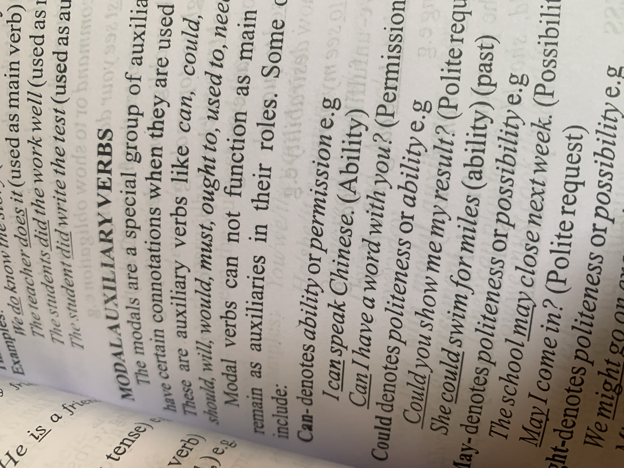 The image is a close-up, angled shot of a page from a book. The text discusses modal auxiliary verbs in English grammar. The page features several examples of modal verbs such as "can," "could," "may," and "might," along with their grammatical functions (e.g., ability, permission, politeness, possibility) and example sentences. The book appears to be open, with text visible on both the left and right pages. The lighting suggests it is an indoor setting, possibly a library or study. The image does not contain any information related to the city of Jauro Yinu, Nigeria.