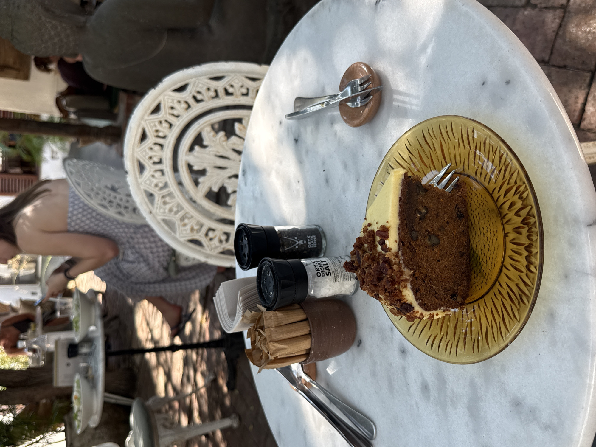 A photograph taken at a high angle shows a round marble table in an outdoor setting, likely a cafe or restaurant, in Cape Town, South Africa. The table is set with a slice of carrot cake on a yellow patterned glass plate, a set of salt and pepper shakers, a small container of sugar packets, silverware, and a small fork rest. A silver fork is embedded in the cake. Two cast-iron garden chairs with ornate patterns surround the table. A woman wearing a light-colored patterned dress sits at an adjacent table. Other patrons are seen in the background. Foliage and sunlight filtering through trees create dappled shadows across the scene. The image captures a casual dining experience.