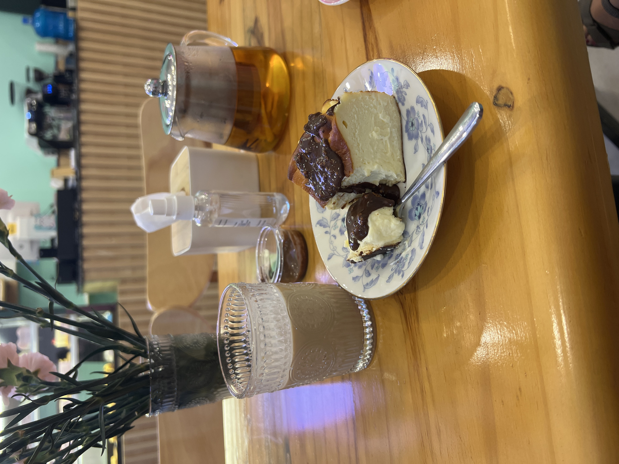 On a wooden table in Bac Nha Trang Ward, Vietnam, sits a teacup filled with a light brown liquid, a glass teapot of amber-colored tea, a bottle of hand sanitizer, a small dish containing a dark brown substance, and a slice of Basque cheesecake with a chocolate topping on a floral patterned plate with a spoon resting on it. A vase of pink carnations is also visible. In the background, a coffee maker and water cooler are discernible on a shelf. A person's foot is visible in the background as well.