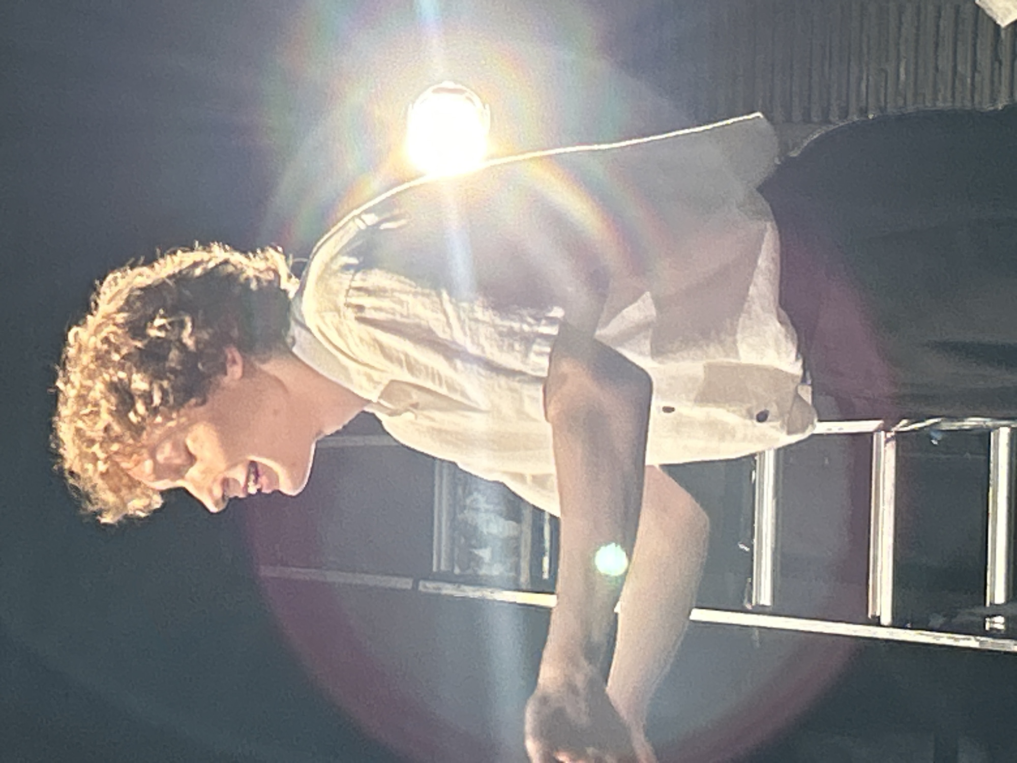 A young person with curly blond hair stands against a dark background with bright lights reflecting on them. They are wearing a light-colored, short-sleeved button-up shirt and dark pants. Their eyes are closed, and their mouth is open as if they are singing or speaking with emotion. Their left arm is extended forward, and their right arm is bent at the elbow, with both hands slightly open. To the left of the person, a silver ladder is partially visible, with rungs extending upwards. The lighting creates lens flares and a rainbow effect on the right side of the image. The overall scene appears to be a performance or a dramatic moment.