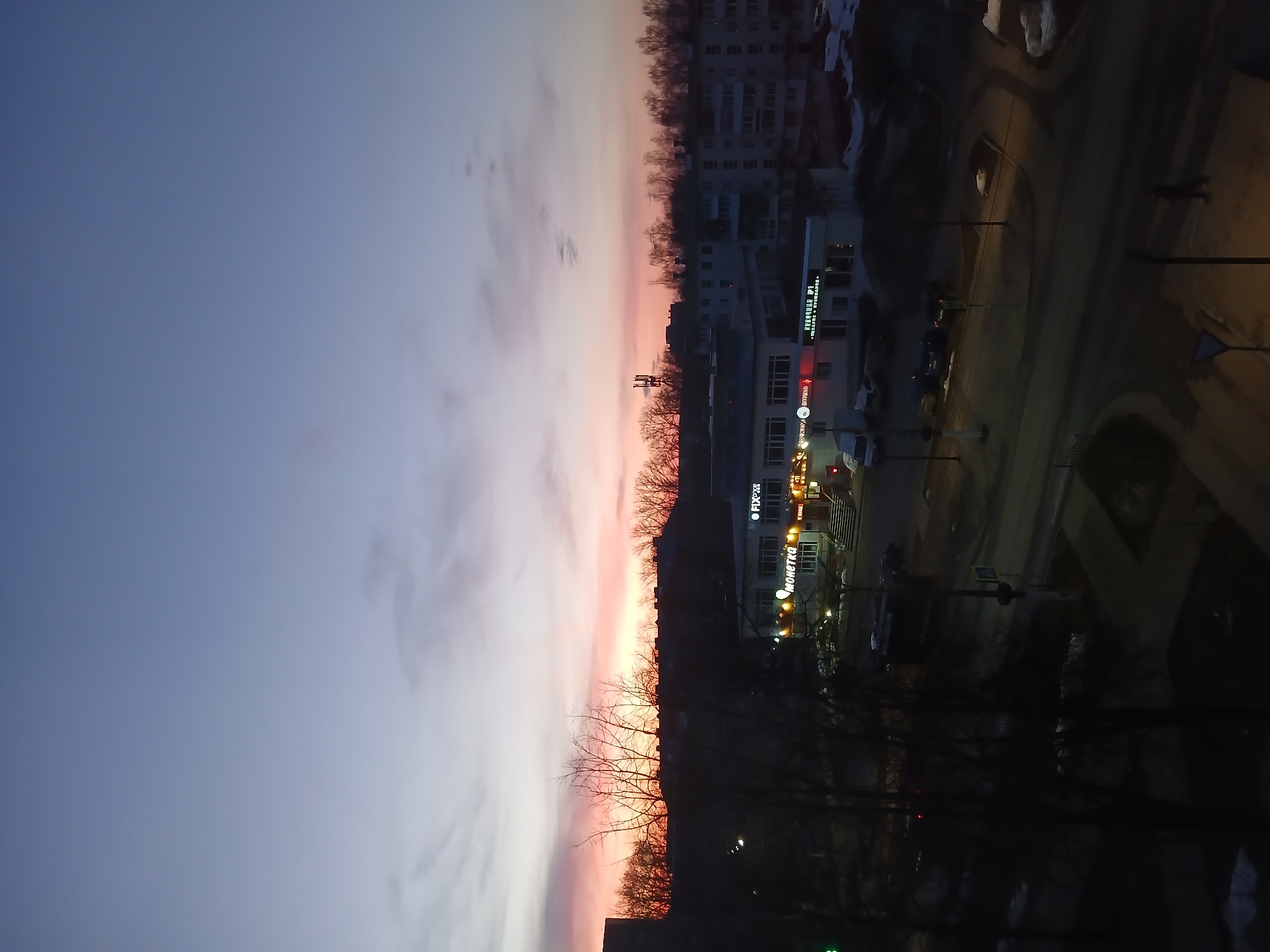 The image depicts a cityscape at dusk or dawn in Chaikovsky, Russia. The sky transitions from blue to pink near the horizon, with scattered clouds. Silhouetted trees are visible along the horizon. Buildings are present on the right side of the image, including a taller residential building and a commercial building with illuminated signs, some in Russian. Streetlights and car headlights are illuminated. A road runs along the right side, showing signs of recent snow or ice. The overall lighting suggests it is early morning or late evening.