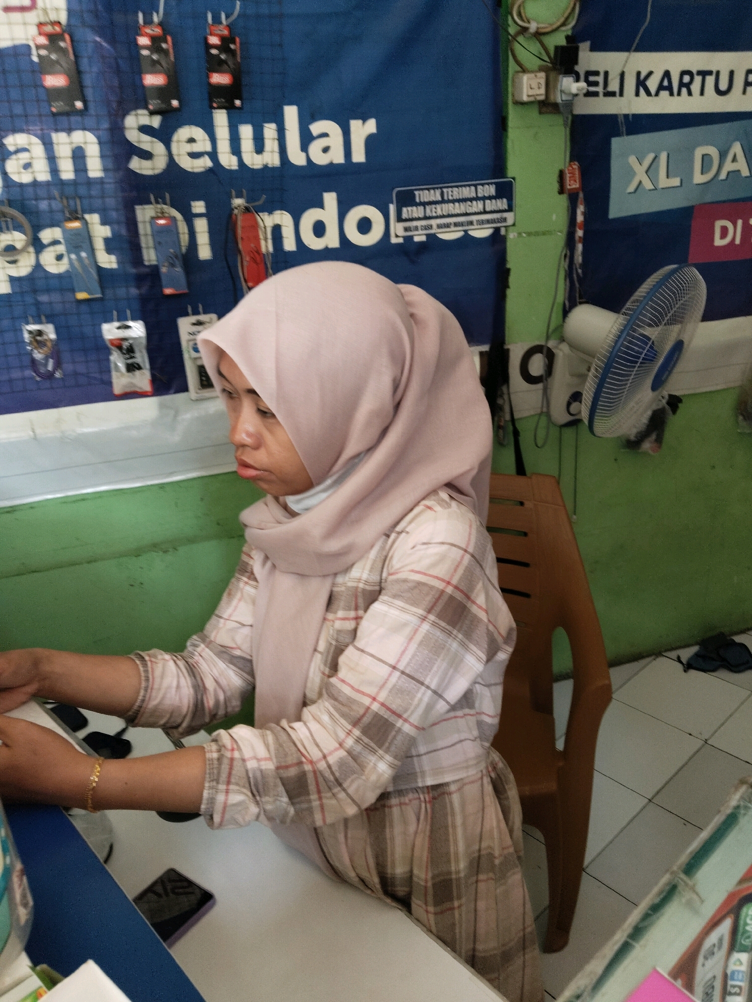 A woman wearing a light pink hijab and a plaid shirt is seated at a desk in what appears to be a small business, likely a cellular phone shop. The background displays blue banners with white text that seems to advertise mobile phone services or products, including "XL DA" and "BELI KARTU PA". Small packaged accessories, possibly phone chargers or earphones, are displayed on a grid-like structure on the wall behind her. A brown plastic chair is positioned to her right. A white electric fan is mounted on the wall to the right of the woman. A mobile phone is visible on the desk in front of her, along with other items on the desk that are partially obscured. The overall environment suggests a retail or service counter setting, likely in Pamanukan, Indonesia.