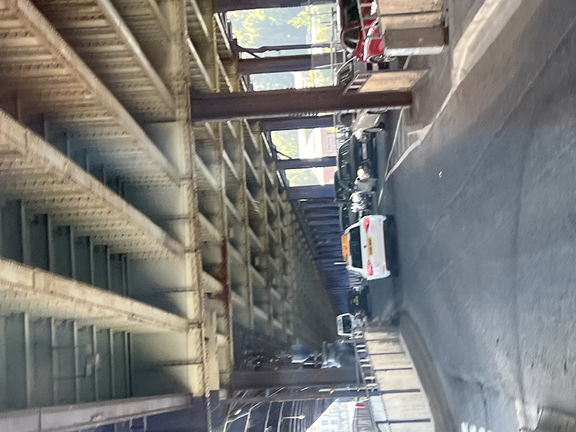 The image shows traffic moving under a large metal bridge structure in the city of Derry/Londonderry, United Kingdom. The bridge appears to be made of steel beams and girders, with a visible underside of latticework and beams. The road below is paved and appears to be a multi-lane route. Several vehicles are visible on the road, including a white car with a yellow sign on its roof, a white van, and several other cars. The environment suggests an urban setting, with the bridge structure dominating the scene. Sunlight is filtering through the gaps in the bridge, creating patterns of light and shadow on the road. There are buildings and trees visible in the distance beyond the bridge.