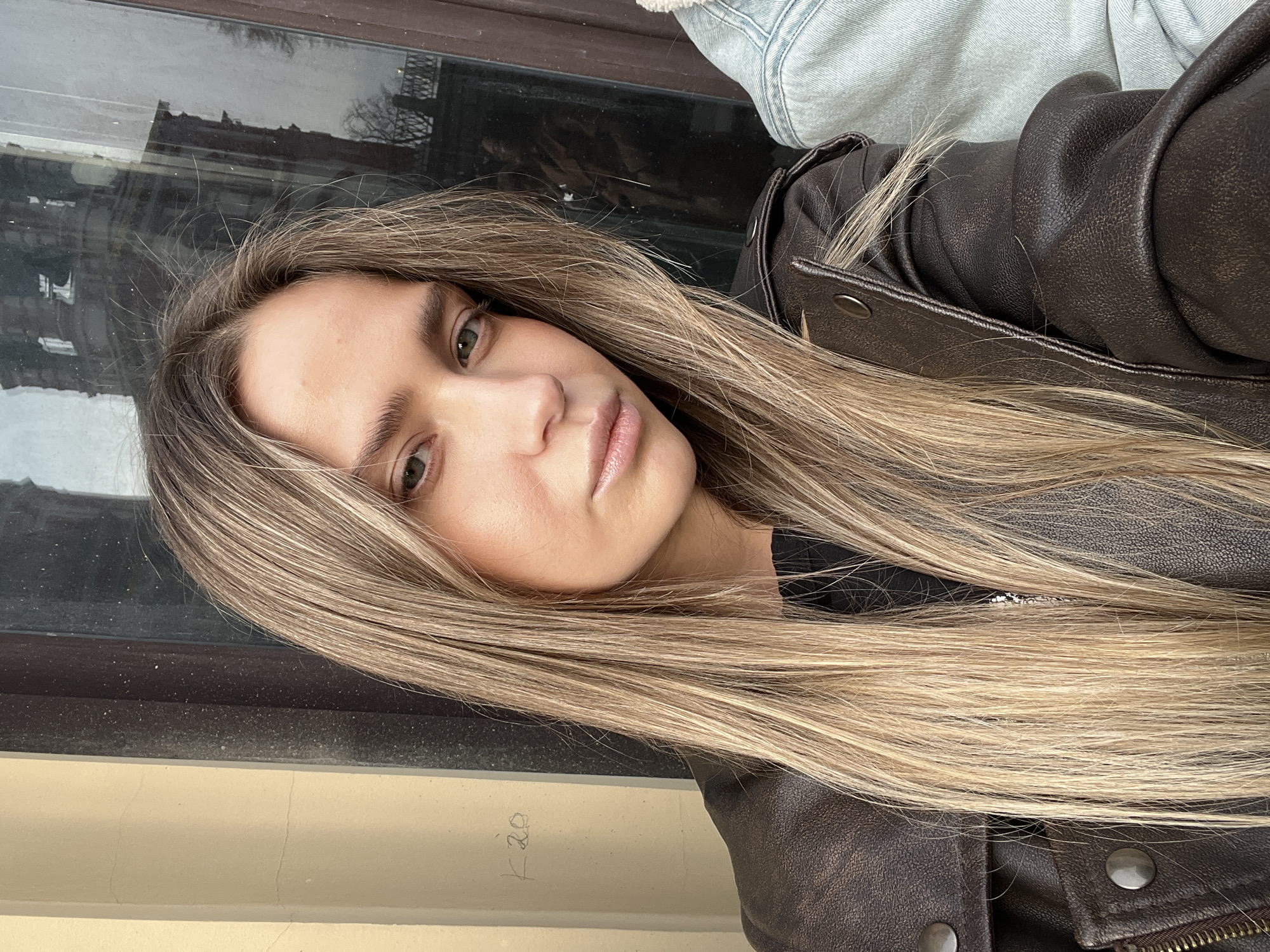 The image is a close-up selfie portrait of a young adult female looking directly at the camera with a neutral expression. She has long, straight, light brown hair with blonde highlights, centrally parted, cascading over her shoulders and chest. Her eyes are green, and her lips are full with a light pink lip product. She is wearing a dark brown leather or faux leather jacket over a black garment, with silver-toned snap buttons visible on the jacket. A portion of a light blue denim garment is visible on the far right, adjacent to the subject.
The background consists of two main elements. To the left, a light beige wall or facade is visible, bearing faint, handwritten characters "K 20." To the right and behind the subject, a dark-framed window reflects an urban outdoor scene. The reflection prominently features a multi-story, dark-colored building with numerous windows and architectural detailing, possibly a historic structure. Bare tree branches are also visible in the reflection, along with a dark metal railing in the lower right. The window surface appears dusty. The specific city of Unknown, Unknown, cannot be determined from the visual evidence; however, the reflections indicate an urban environment with older architecture.