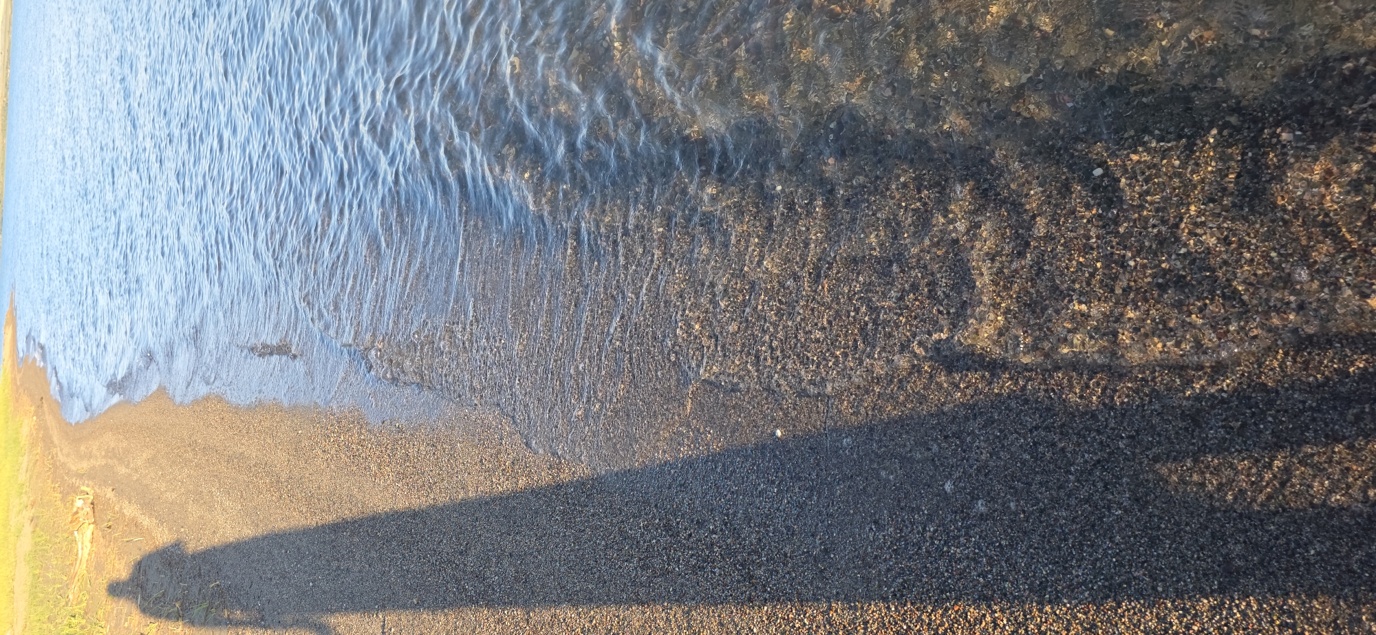 The image depicts a sunlit shoreline on a body of water, likely Issyk-Kul Lake in Kyrgyzstan, given the distinct light and vegetation visible on the upper left. The foreground shows a narrow strip of dark, fine gravel beach. A long, distinct shadow of a person, presumably standing on the beach, stretches across the gravel and into the shallow water. The water itself is clear, revealing the pebbly lakebed beneath the surface. Gentle waves lap at the shore, creating ripples and a shimmering effect on the water. The background is dominated by the tranquil blue expanse of the lake, meeting a pale blue sky at the horizon. The overall impression is one of peaceful solitude and the natural beauty of a lakeside environment.