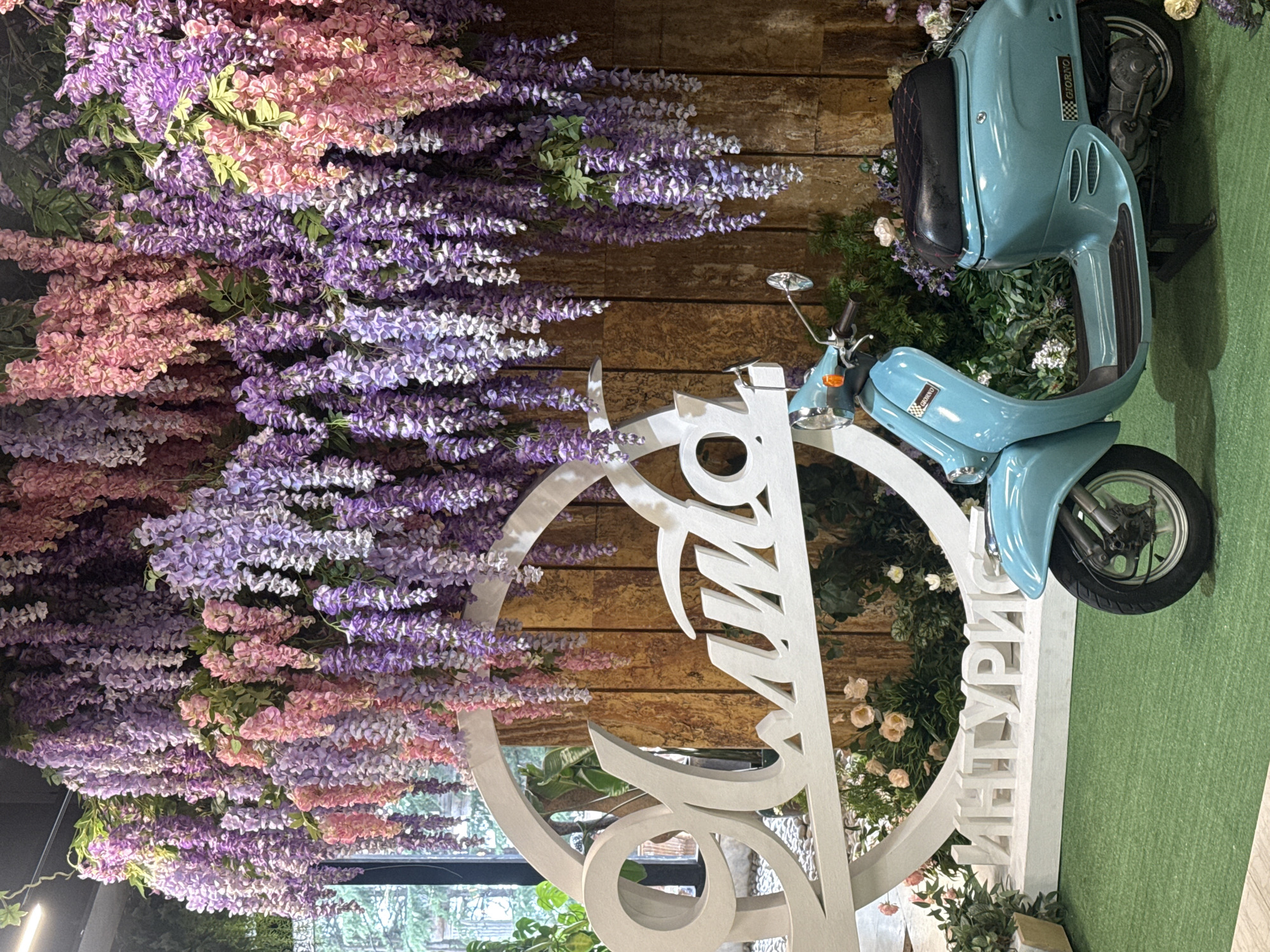 The image depicts a decorative display featuring a teal-colored scooter placed in front of a wooden-paneled wall adorned with cascading artificial wisteria in shades of pink and purple. A large white circular frame with stylized lettering, appearing to read "ЯЛТА" (Yalta) and "ИНТУРИС" (Intourist), is situated behind the scooter. The ground is covered with green artificial turf, and there are some artificial plants and flowers interspersed around the base of the display. The scene is likely set up as a photo opportunity.
