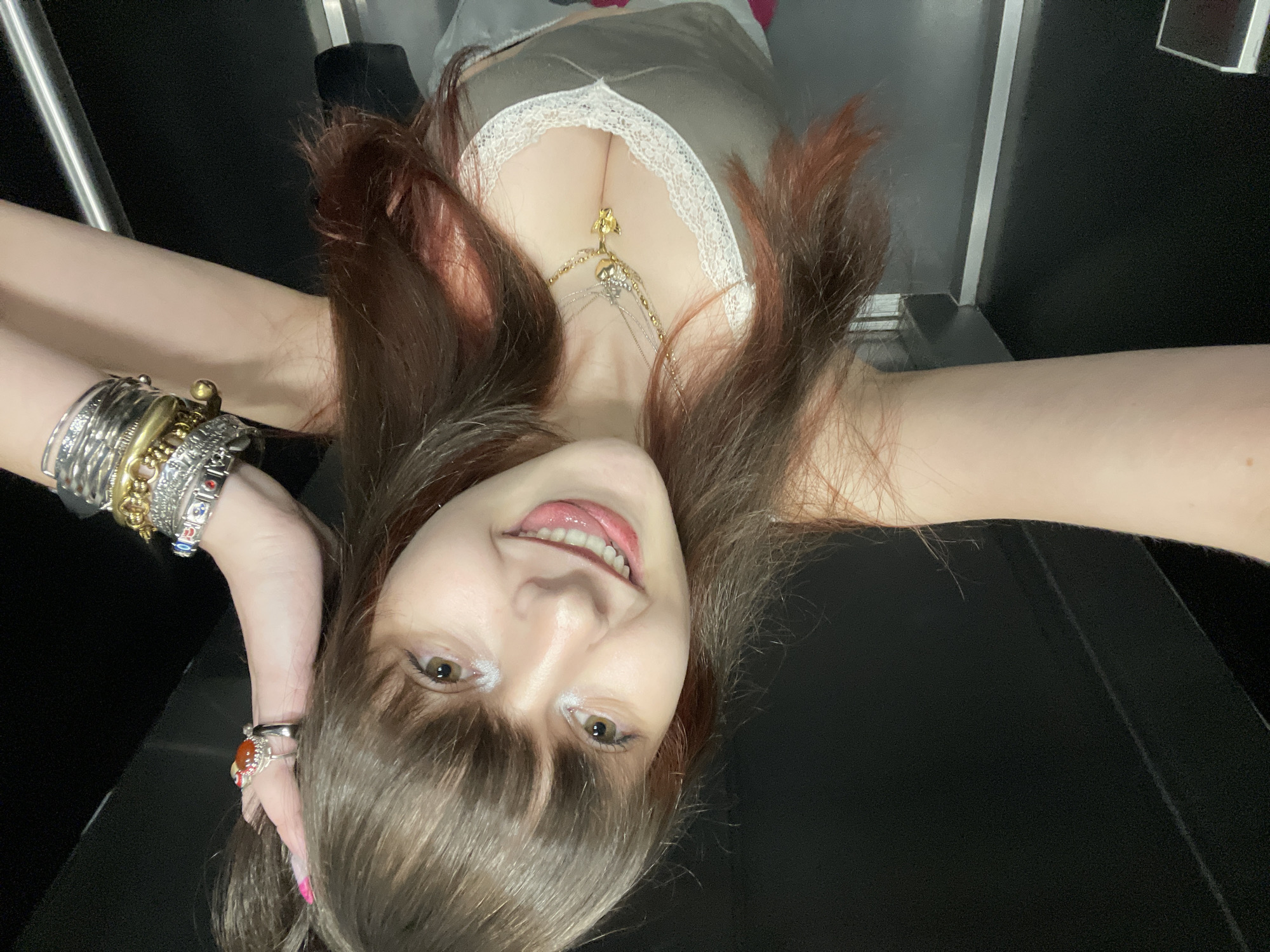 A woman lies on a dark surface, possibly a bench or seat, oriented at an angle to the camera. She wears a low-cut, light green top with white lace trim and multiple necklaces, including one with a gold pendant. Her brown hair has reddish highlights, and she has fair skin, wearing makeup. She is smiling slightly with her tongue visible. A stack of bracelets adorns her left wrist. The background consists of a light gray wall with a metallic accent and a dark, likely black surface. The location is Raanana, Israel, but the photo does not provide information to be more specific within the city. The image is a selfie.