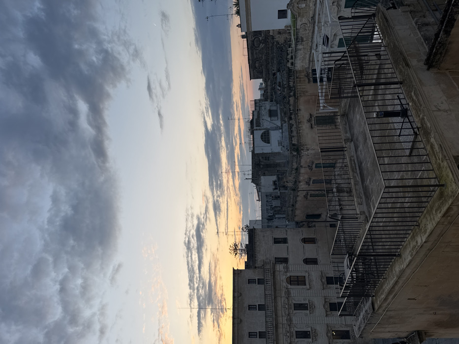 The image depicts an urban landscape in Lecce, Italy, observed from an elevated perspective, likely a rooftop, during either sunrise or sunset. The sky, partially covered by scattered grey and white clouds, exhibits a warm gradient from orange and yellow hues near the horizon on the left to lighter blue and grey tones overhead and towards the right. A faint crescent moon is visible in the upper right quadrant of the sky.
In the immediate foreground, a flat rooftop terrace is enclosed by dark, vertical metal railings. The terrace floor is paved with light-colored tiles. On the left section of this terrace, a dark metal table and two chairs are present. Towards the right, a dark, cylindrical object, possibly a parasol base or ventilation fixture, rests on a square base on the terrace floor.
The midground is dominated by a dense arrangement of multi-story buildings, typical of historic Italian urban architecture. A prominent, light-colored building on the far left features multiple rectangular windows, arched doorways, and ornate classical elements on its facade. Behind and to the right of this structure, numerous other buildings of varying heights and ages are visible, characterized by their weathered facades in shades of beige, brown, and grey. Many rooftops are equipped with an array of television antennas and other communication aerials. A tall, slender conifer tree rises above one of the central buildings.
The background shows additional buildings receding into the distance under the fading or emerging light, with the overall scene illuminated by the indirect glow of the sun. There are no individuals present or engaged in specific actions within the frame.