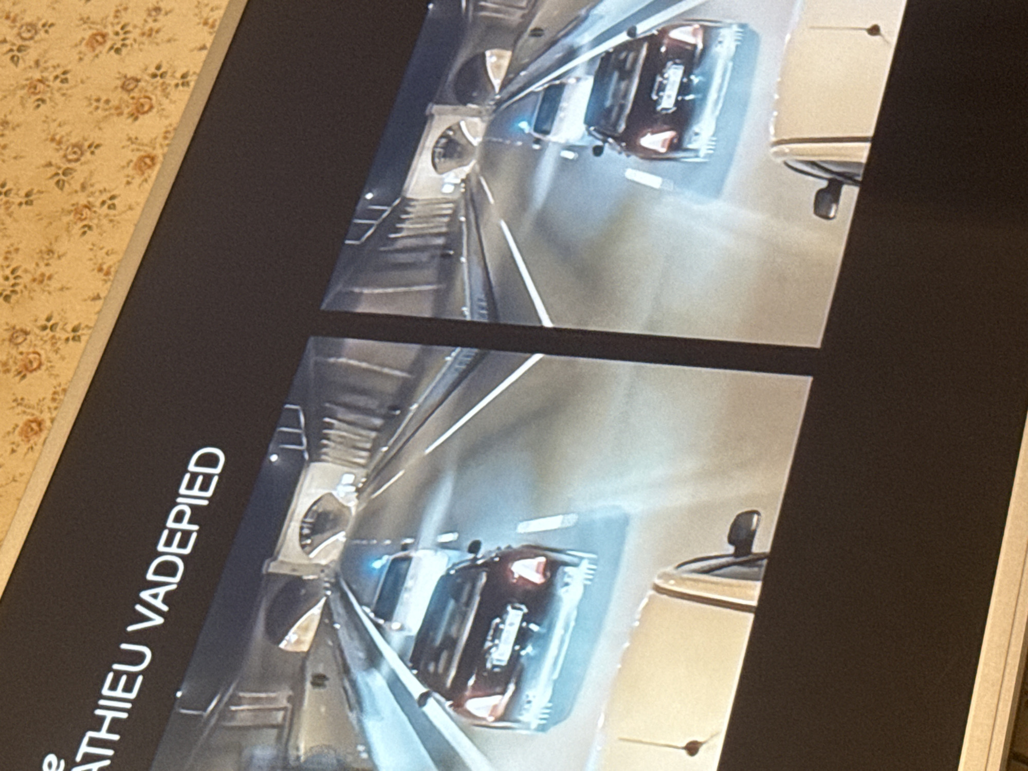 The image shows a framed photograph or poster displaying a perspective view of a tunnel with cars. The tunnel appears to be well-lit with artificial lighting. Two cars are visible, one dark-colored and one light-colored, traveling inside the tunnel. The name "MATHIEU VADEPIED" is written on the frame or border of the photograph. The image is taken indoors, as indicated by the floral wallpaper visible in the background. The location of the tunnel and the city of Mytishchi, Russia, are not discernible from the image.