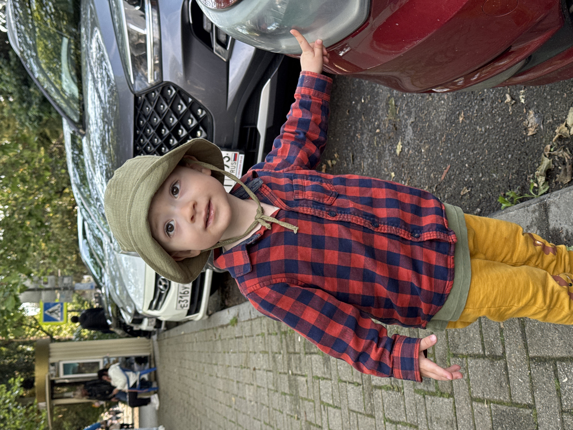A toddler in a green hat and red and blue plaid shirt stands on a paved sidewalk next to several parked cars. The child is wearing yellow pants and is touching the headlight of a red car with their right index finger. To the left of the red car, there is a gray car with a Russian license plate. Further to the left, a white car is parked. In the background, a building and trees are visible, and a pedestrian crossing sign can be seen. People are visible in the background near a building. The license plate on the gray car indicates that this image was taken in Sochi, Russia.