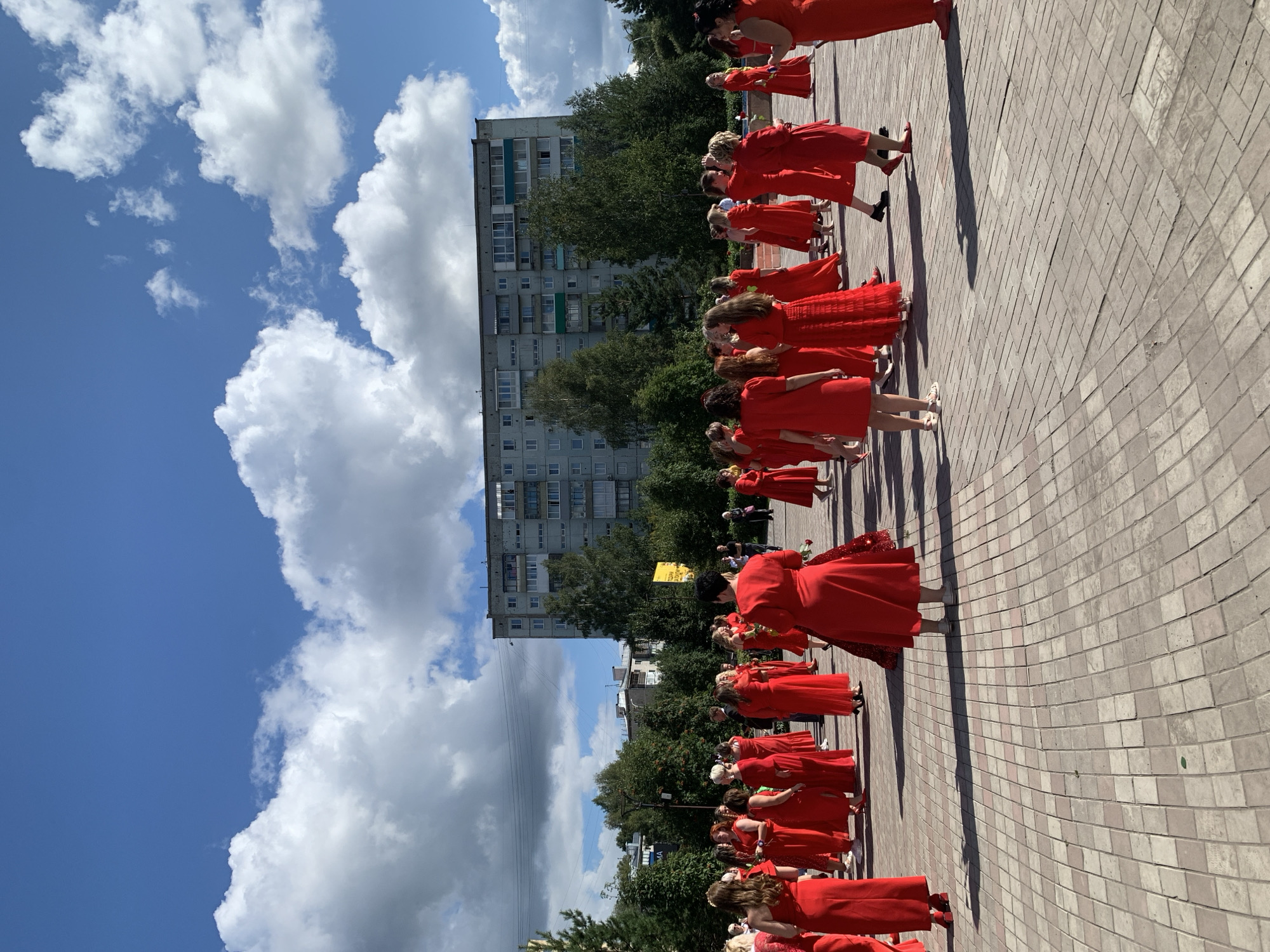In Prokopyevsk, Russia, a group of women dressed in matching red dresses are gathered on a paved outdoor area. The women are standing in various formations and appear to be participating in an event or performance. Behind them, a large, multi-story residential building dominates the skyline, with trees and a bright blue sky filled with fluffy white clouds. The ground is paved with light-colored square tiles, and shadows from the women and trees are cast across the surface. Some of the women are holding red roses, suggesting a celebratory or ceremonial occasion. The scene captures a moment of communal activity in an urban setting.