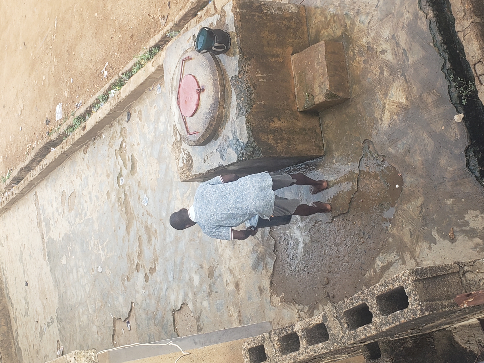 Location: Alalubosa, Nigeria.
Scene: A man stands on a damp, cracked concrete surface. The surface is partially covered with water and debris. The man is facing a circular, metal well cover with a red top. A black bucket rests on the well cover. The man is wearing a light blue shirt, dark shorts, and sandals. He appears to be washing his hands, or something held in his hands. A concrete block wall is partially visible in the lower right corner of the image. A narrow ditch runs along the right edge of the concrete surface. Additional concrete surfaces are seen in the background. There is dirt and some scattered vegetation on the left.