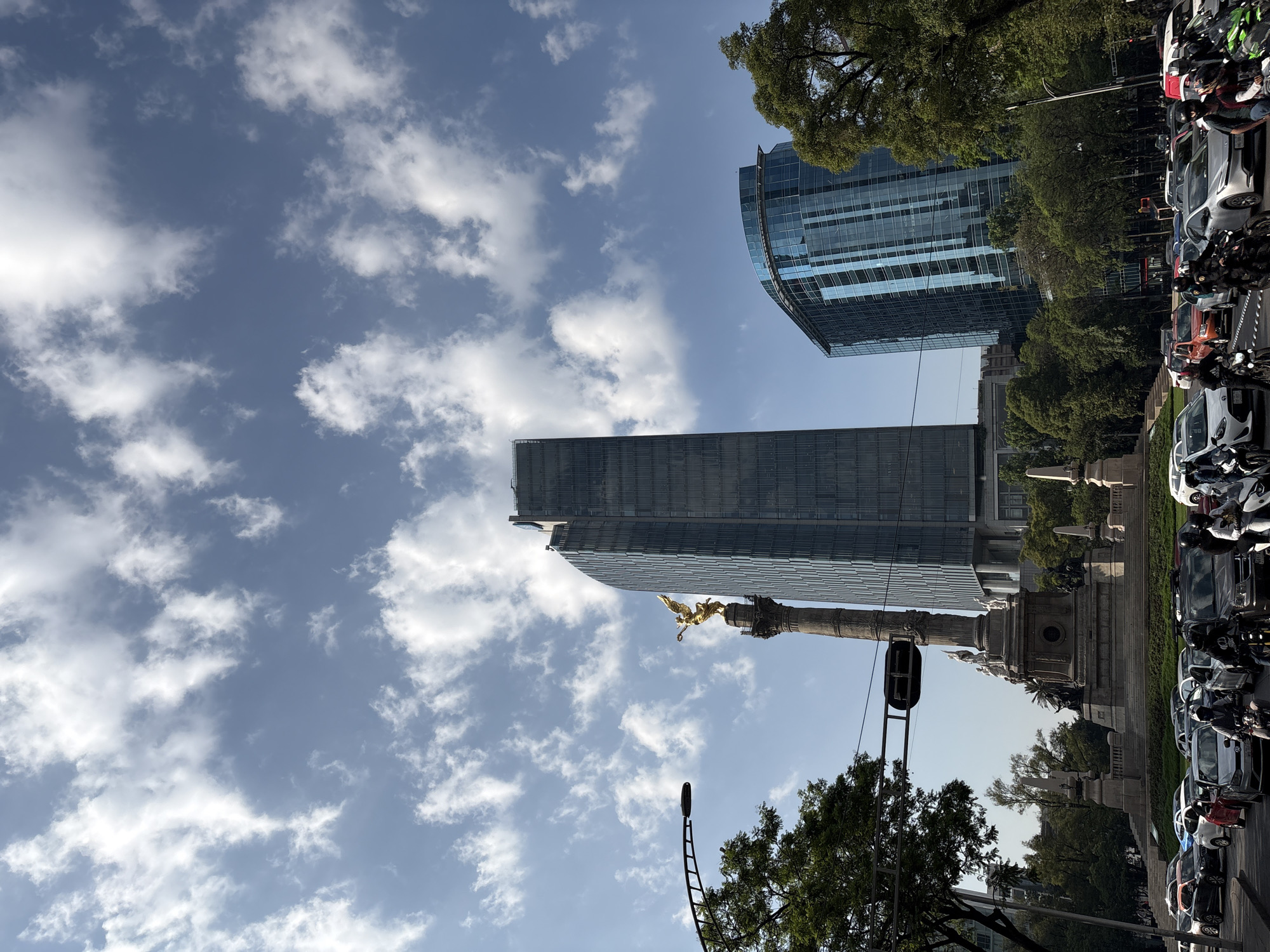 The image captures the iconic Angel of Independence monument in Mexico City, Mexico, with a low-angle perspective. The golden angel statue stands atop a tall column, and modern skyscrapers rise in the background against a blue sky with scattered clouds. A traffic light and a street lamp are visible near the monument. On the right side of the frame, there is a row of parked cars and motorcycles along the street, partially obscured by trees. The environment includes clear daytime conditions and elements of urban infrastructure.