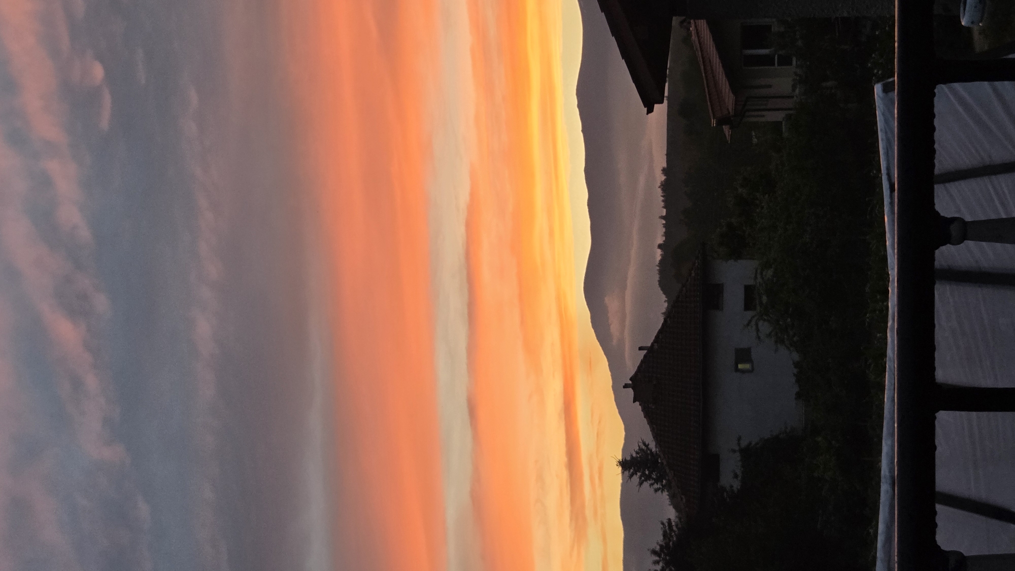 A vertical, eye-level shot depicts a dramatic sunset over a village. The sky is a vibrant gradient of colors, transitioning from a dusky gray and pink at the top to fiery orange and yellow closer to the horizon. Wispy clouds are scattered across the upper portion of the sky. Below the sky, silhouetted mountain ranges are visible, partially obscured by mist. In the foreground, the rooftops of houses peek through dark green foliage. A white house with a red-tiled roof is prominent on the left, with several windows visible. Another house with a red-tiled roof is visible on the right. A dark wooden railing runs along the bottom of the frame, obscuring the lower part of the scene, which appears to be a patio or balcony. The overall atmosphere is serene and picturesque.