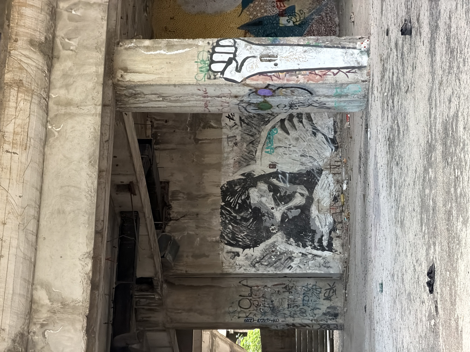 The image depicts the interior of an abandoned, unfinished concrete structure in Mostar, Bosnia and Herzegovina. The space is characterized by exposed concrete columns, beams, and unfinished ceilings, with signs of water damage and deterioration. The ground is dusty and littered with debris.
Dominating the scene is a large black and white mural of a person's face, rendered in a stylized, somewhat somber manner, painted on a concrete wall. To the right of the mural, on a concrete column, is a stencil of a raised fist, a common symbol of protest and solidarity. The column itself is covered in graffiti in various colors and languages.
Graffiti is also present on other walls and surfaces, with tags and inscriptions visible throughout the frame. In the background, stairs can be seen leading upwards, suggesting multiple levels to the structure. Sunlight streams in from an opening on the left, illuminating parts of the scene and casting shadows.
The overall impression is one of decay and artistic intervention in a forgotten urban space. There are no people visible in the image. Scattered on the floor are a few discarded items, including what appears to be a pair of dark fabric shoes or a bundle of clothing in the foreground, and a single dark shoe further back. A small, white stuffed toy is visible near the base of the column with the raised fist.