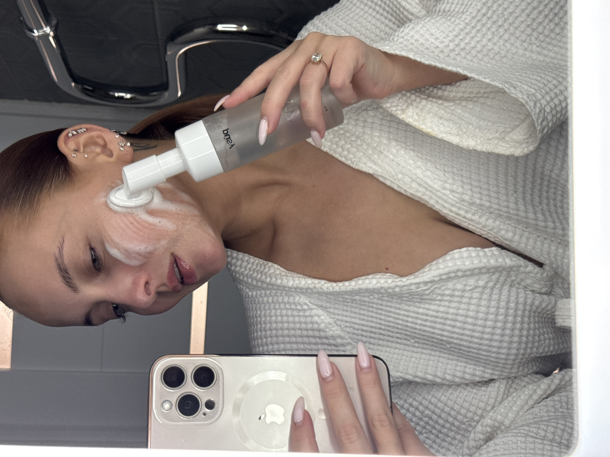 A woman in a white waffle-textured bathrobe is in a bathroom. She is applying a white foaming cleanser from a clear bottle with a white pump to her face, while holding a white smartphone (iPhone) with multiple camera lenses in her other hand. The woman has brown hair pulled back, fair skin, and is wearing multiple earrings in her left ear. Her fingernails are long and painted pale pink. She is wearing a ring on her finger of the hand holding the bottle of cleanser. A chrome faucet is visible in the background. The location is likely a residential bathroom, the city of Chaikovsky, Russia.