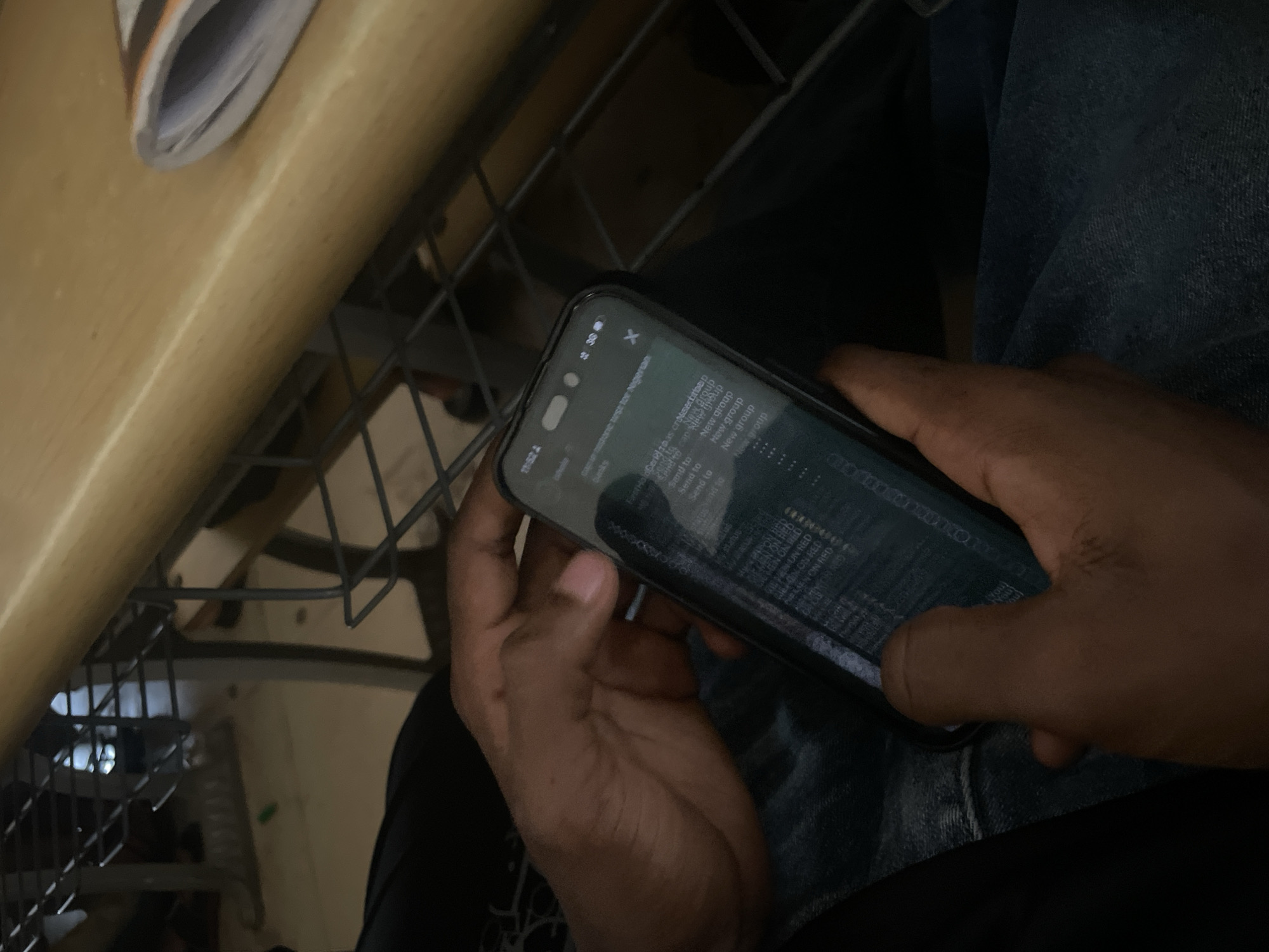 A person is holding a smartphone in their hands. The phone screen displays a green interface with text. The phone is held in front of the person's lap, and their hands are positioned to operate the device. The environment appears to be indoors, possibly a classroom or office, with a wooden table and metal grating visible in the background. The overall lighting suggests an indoor setting.