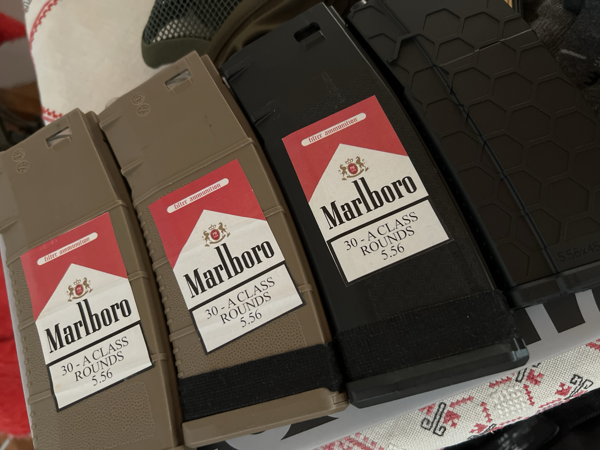 Four firearm magazines are arranged closely together on a surface, likely indoors. Two magazines on the left are tan/brown, and two on the right are black. Each magazine features a custom label designed to mimic the classic Marlboro cigarette packaging.
The labels prominently display the word "Marlboro" in the brand's signature font, beneath a red and white crest that includes the letters "PM" and a crown. Below the Marlboro branding, the labels read "30 - A CLASS ROUNDS 5.56". Above the main Marlboro logo, a small banner on each label states "filter ammunition".
The tan magazines on the left have small circular markings on their upper surfaces. A black elastic or fabric band is wrapped around the lower portion of the second tan magazine and the first black magazine. The black magazines on the right display a subtle hexagonal textured pattern on their sides. Faintly visible on the side of the far-right black magazine is the text "5.56x45". The magazines are resting on a white fabric surface with a recurring red embroidered pattern, possibly a tablecloth or blanket. A dark, textured object and another dark, smooth object are partially visible in the background.