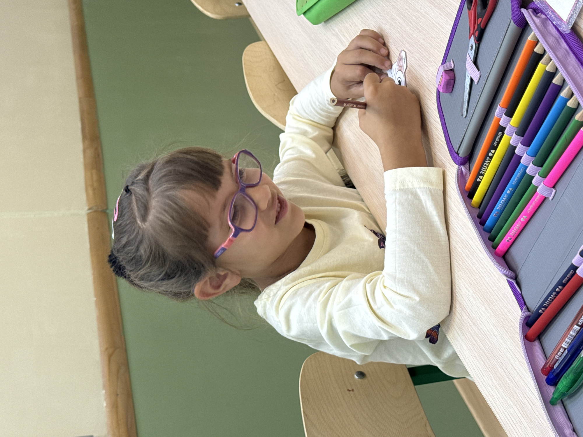 A young girl with dark hair pulled back and pink-rimmed glasses is seated at a light wood desk, actively engaged in drawing or coloring. She is wearing a long-sleeved cream-colored shirt. Her right hand holds a brown colored pencil, which she is using to color a small cutout figure on the desk. In front of her is an open purple pencil case filled with a variety of colored pencils, a small pair of scissors, and a pink eraser. The background consists of a light beige wall with a dark wood trim and a muted green wall below. The image appears to be taken indoors, possibly in a classroom or a dedicated study area. There is no identifiable information within the image to suggest a specific location in the city of Perm, Russia.