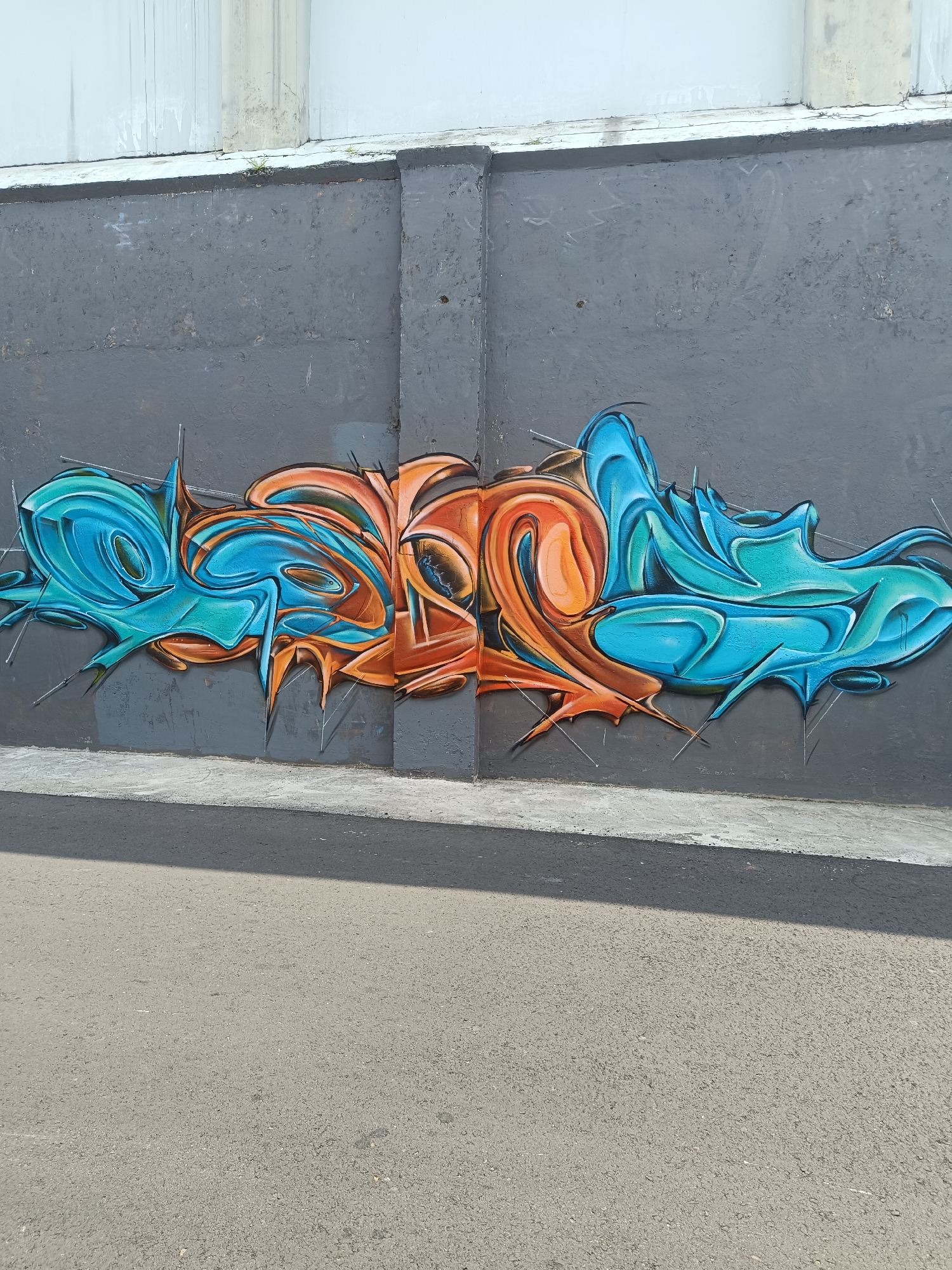 A graffiti artwork is displayed on a weathered, dark gray concrete wall. The artwork is abstract and features bold, flowing shapes in contrasting shades of bright turquoise blue and burnt orange. The turquoise sections appear on the left and right sides of the piece, while the orange dominates the center. The graffiti has a three-dimensional, bubble-like quality with sharp edges and highlights that suggest light reflecting off its surface. Thin, silver wires or metal supports are visible extending from various points of the graffiti into the wall, giving the impression that the artwork is suspended or attached to the surface. The wall itself is textured and shows signs of age, with faint marks and a lighter gray stripe running horizontally across the lower portion. Above the graffiti, the upper section of the wall is lighter and appears to be a different material, possibly concrete, with vertical streaks of discoloration. The ground in front of the wall is an asphalt road, with a distinct shadow cast across it from an unseen object to the left, suggesting the presence of sunlight. The scene appears to be an outdoor urban environment. The graffiti's style and placement are consistent with street art often found in public spaces in cities like Yogyakarta, Indonesia, though the specific location cannot be definitively identified without further context.