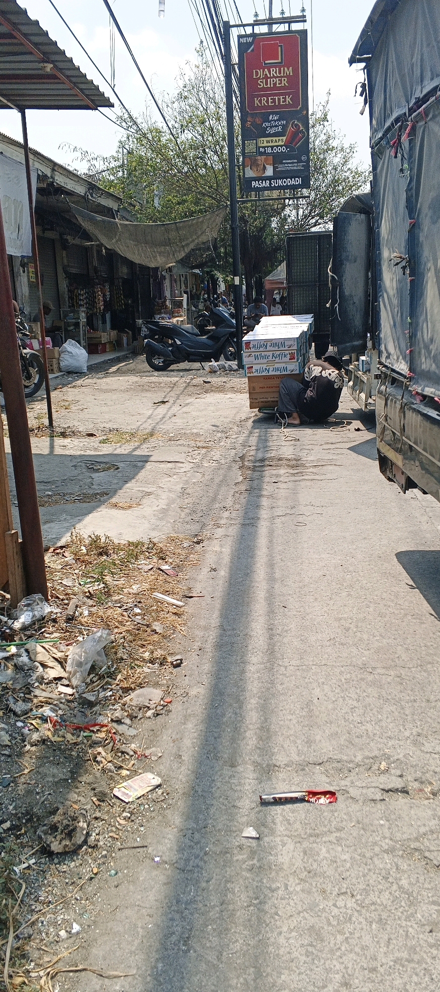 The image depicts a street scene in Sukodadi, Indonesia. The foreground shows a weathered concrete road with scattered debris, including dry leaves, plastic bags, and what appear to be broken pieces of packaging and a used cigarette pack. In the middle ground, a dark-colored motorcycle is parked, and to the right, several stacks of white boxes labeled "White Koffie" are piled on the ground. A person wearing a dark shirt and grey pants is seated on the ground, leaning against the boxes. Behind these boxes, the side of a large dark-colored truck is visible. Further in the background, on the left side of the image, a row of market stalls with covered awnings lines the street. A prominent vertical sign on a pole displays an advertisement for "DJARUM SUPER KRETEK" cigarettes, mentioning "PASAR SUKODADI." The overall impression is of a busy, possibly commercial area with elements of everyday life and some degree of litter. The lighting suggests it is daytime, with shadows cast across the road.