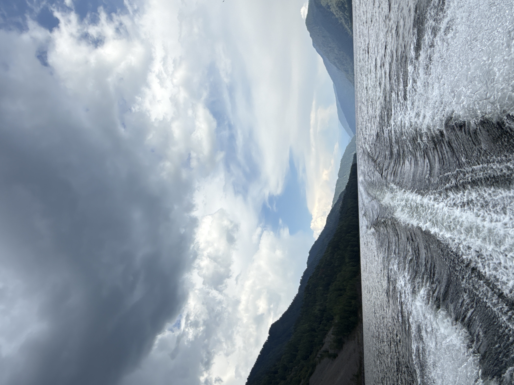 The image depicts a wide view of a lake from the perspective of a moving boat. In the foreground, the boat's wake churns the dark water, creating white foam and intricate wave patterns. The lake stretches towards the horizon, bordered by dense, dark green forests covering the steep slopes of mountains on either side. The sky above is a dramatic mix of dark, brooding storm clouds and patches of bright blue sky with scattered white clouds. The overall impression is one of movement and a vast, natural landscape under a dynamic sky. The location is Kordon Baigazan, Russia.