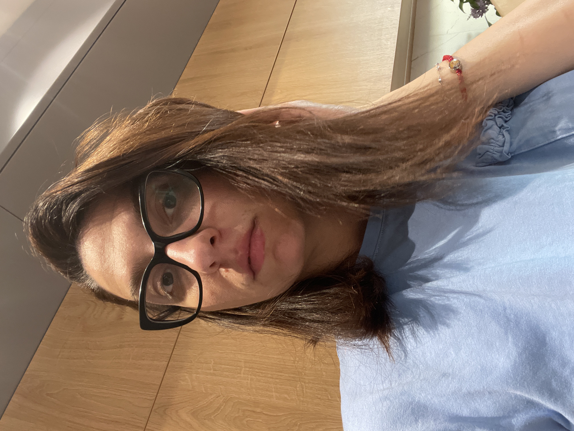 A woman with dark hair and glasses is indoors, possibly in an apartment or house. She is wearing a light blue shirt. A red bracelet is on her wrist. The background consists of wooden paneling and a white wall. The scene appears to be indoors, likely a residential setting. There is no information to suggest that the image was taken in Sofia, Bulgaria.