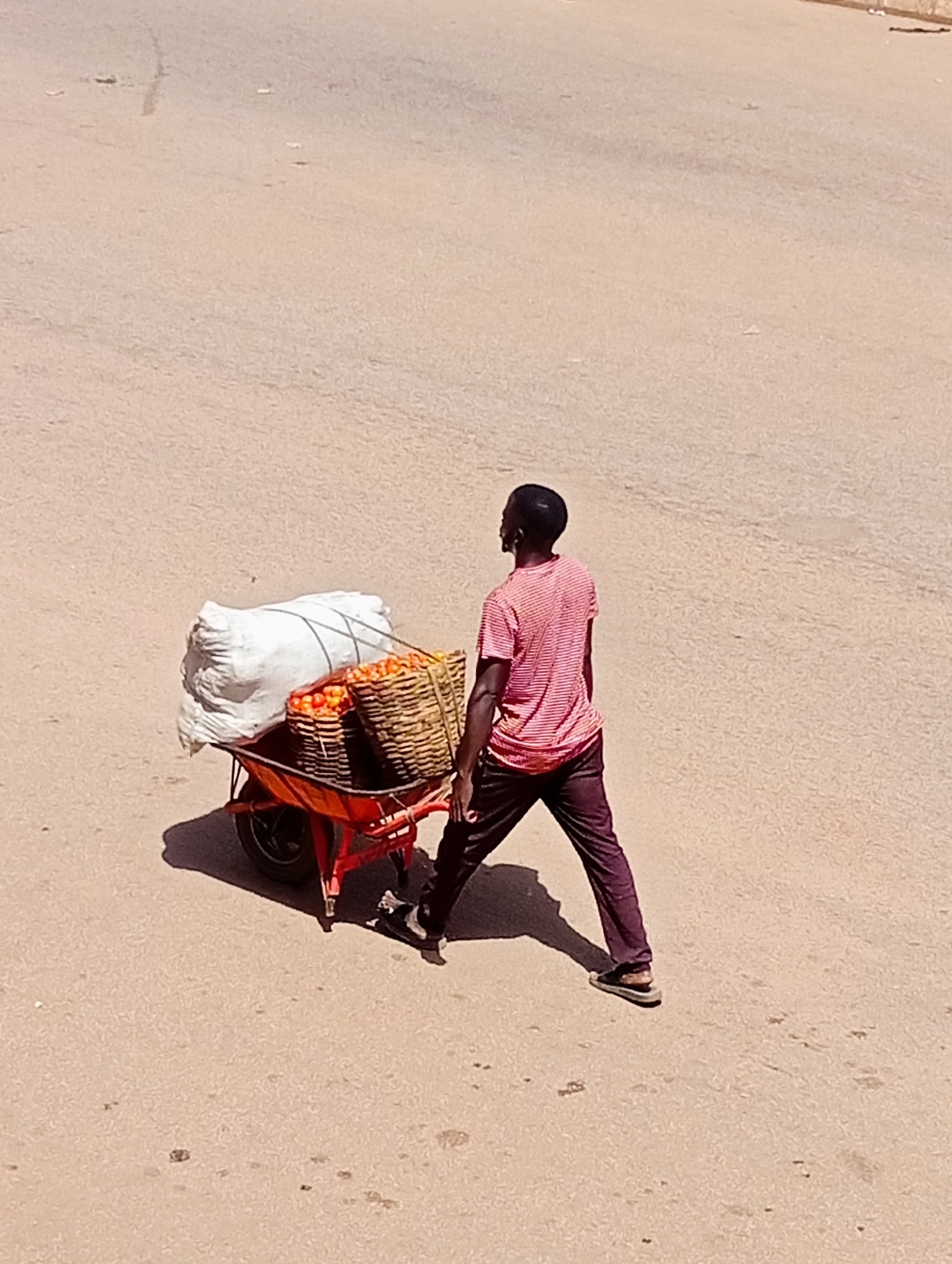 A man wearing a red and white striped t-shirt and dark pants is pushing a red wheelbarrow loaded with two woven baskets of tomatoes and a large white sack. The man is walking on a paved road in Bukuru, Nigeria, with the sun casting a shadow from the wheelbarrow.