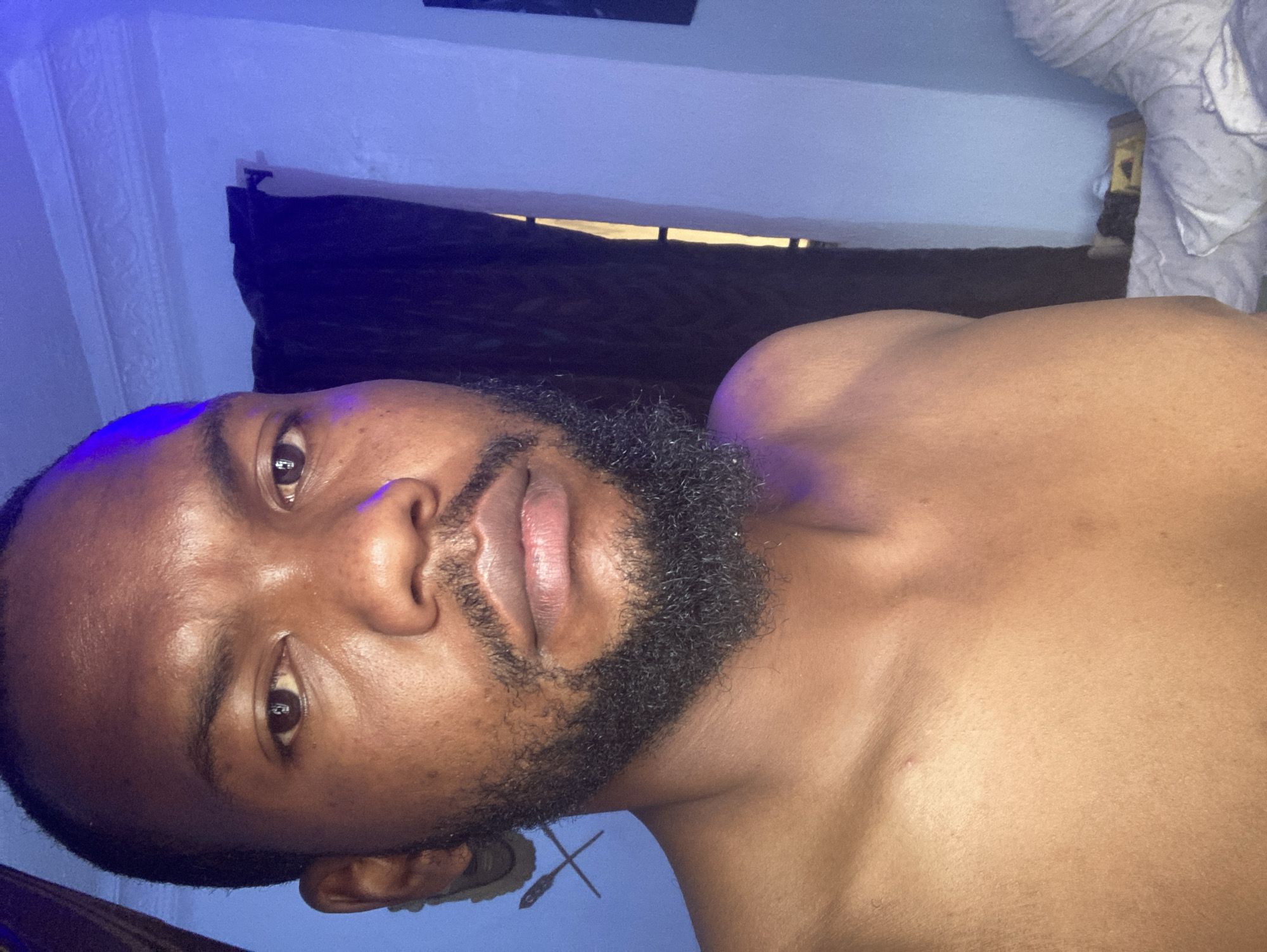 A close-up, low-angle shot reveals a dark-skinned man with a closely cropped beard, lying on what appears to be a bed. He has brown eyes and a serious expression. The room is illuminated with blue-toned lighting. Behind him, a window is partially obscured by dark curtains. A crumpled white sheet is visible in the top right corner. The location in the city of Jos, Nigeria cannot be determined from the image.