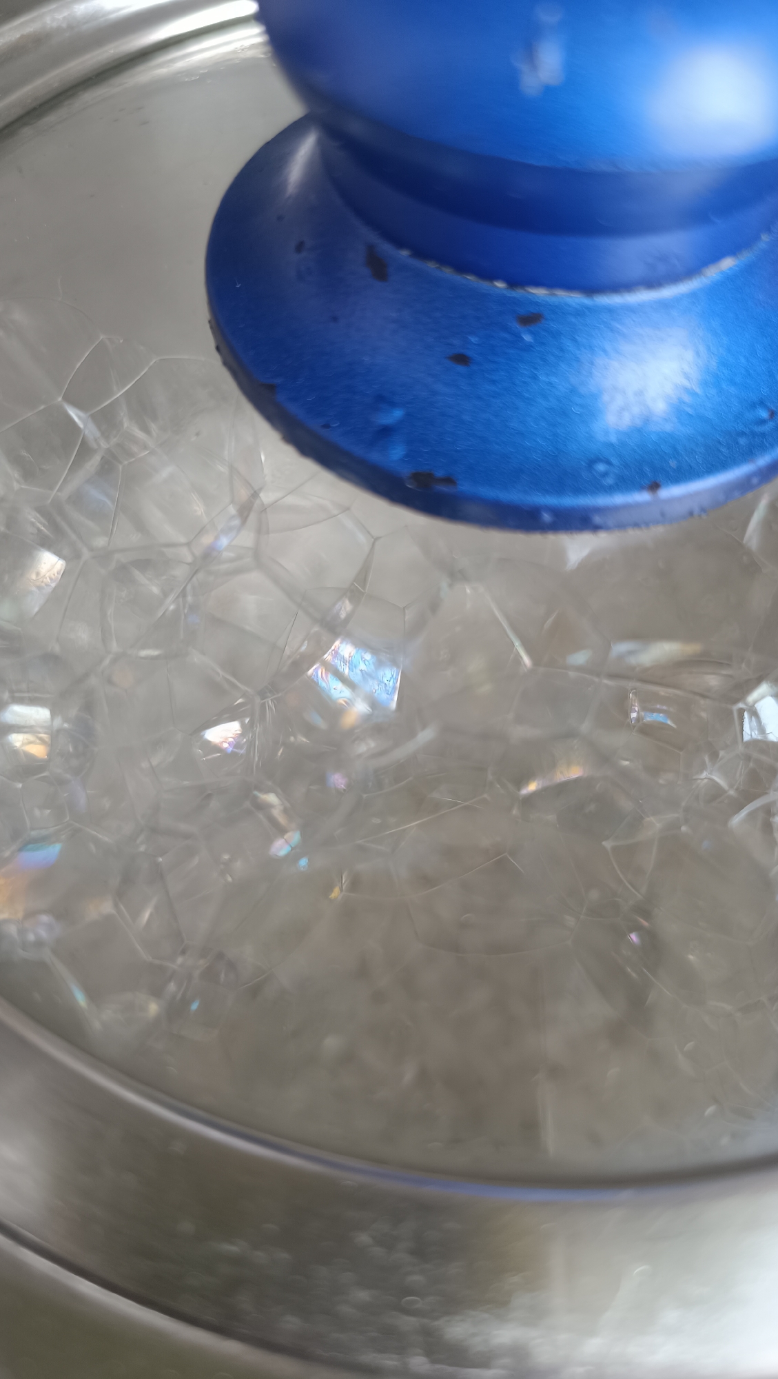 A close-up, high-angle shot shows a pot with a clear glass lid topped by a blue knob. The pot appears to contain soapy water, evidenced by a dense formation of iridescent, translucent bubbles visible through the lid. The bubbles are of varying sizes and shapes, closely packed together, with some reflecting rainbow-like colors. The blue knob has visible imperfections, with some darker smudges or spots along its edges. The pot's rim is partially visible, appearing to be metallic. No people, animals, or specific environmental cues tie the image to Lermontov, Russia; the content is strictly limited to the pot with soapy water.